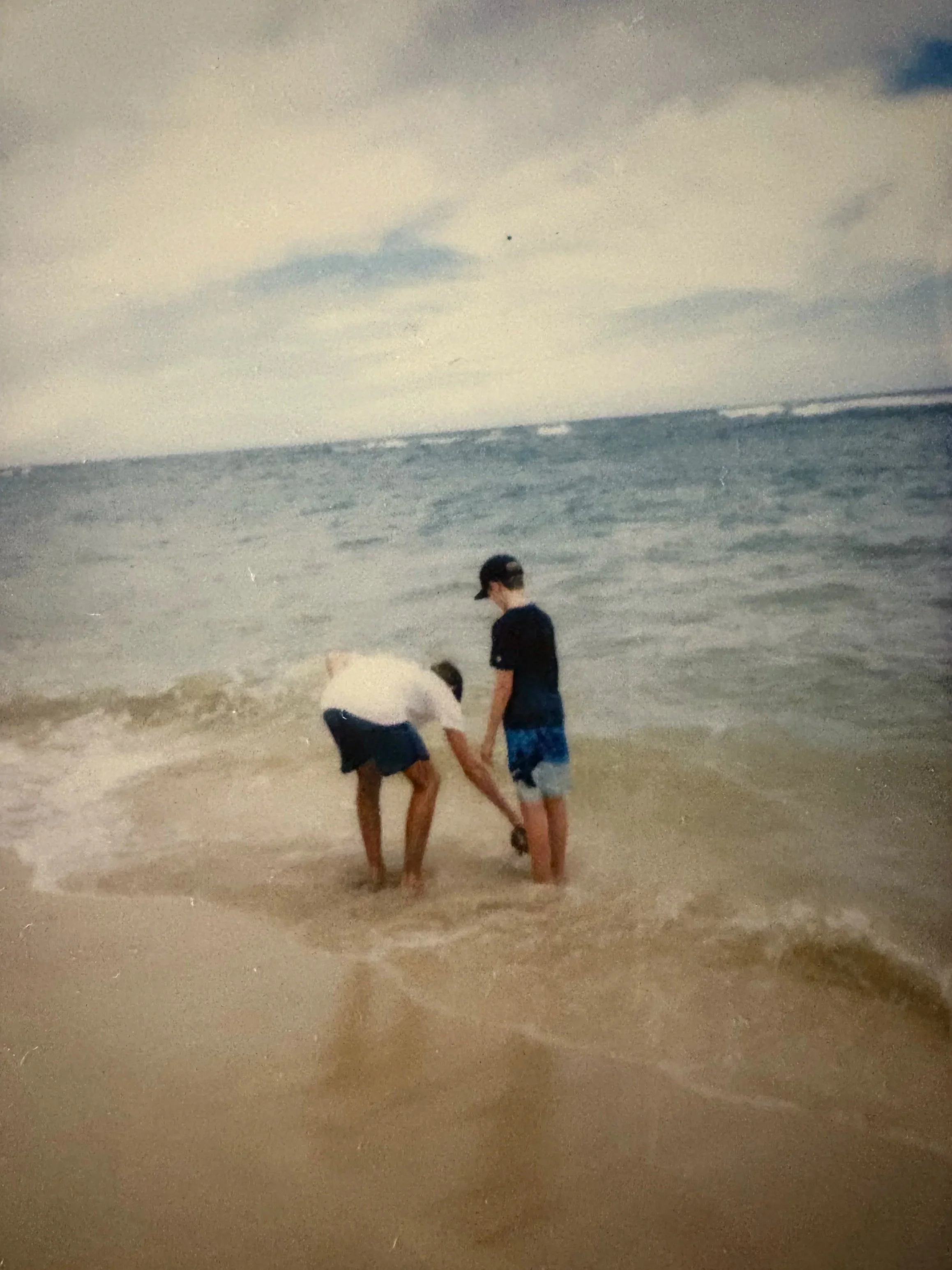 Photograph taken by Aleka London featuring two boys playing in shallow ocean water at the beach under a cloudy sky, the ocean waves gently splashing over their ankles.