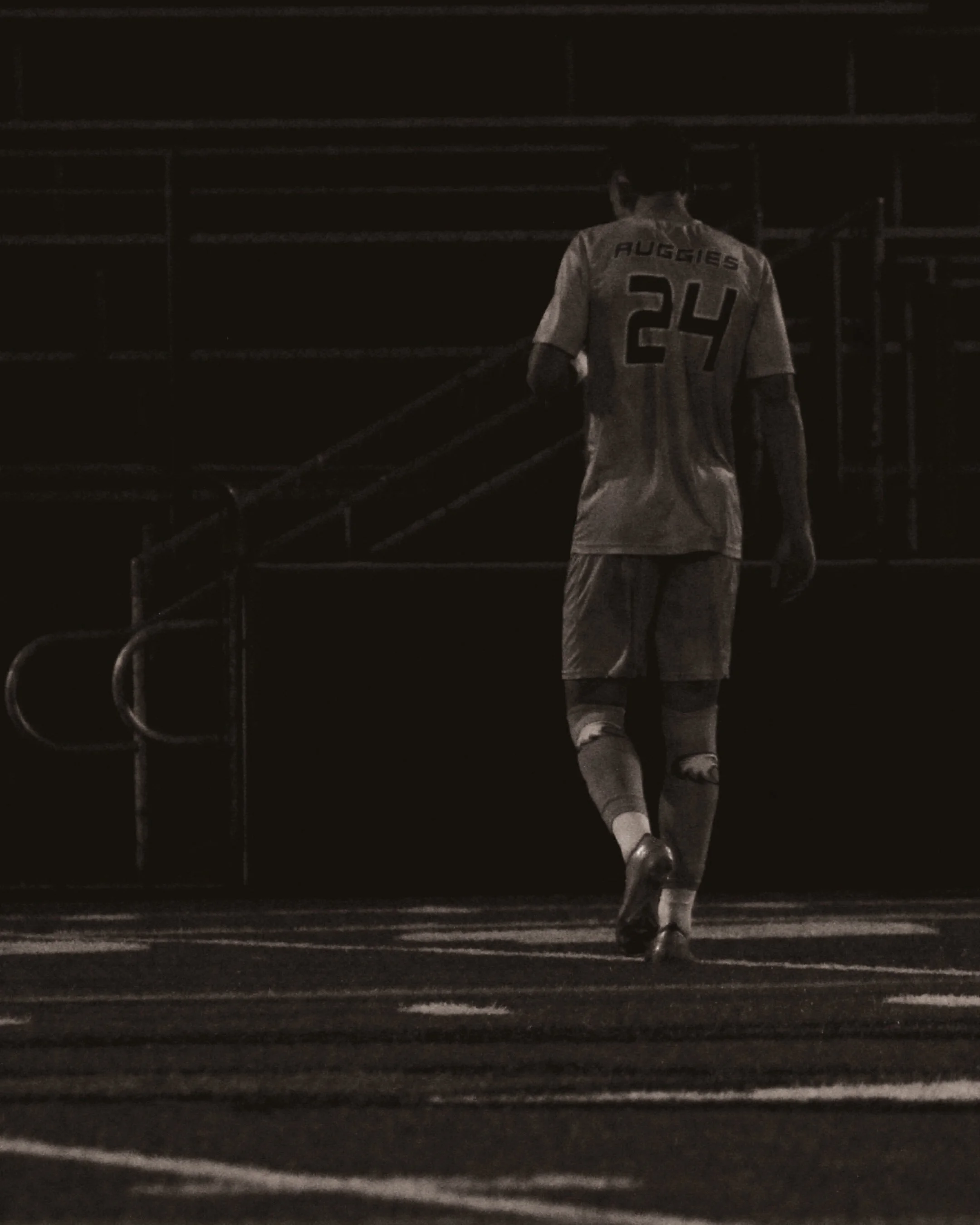 A soccer player wearing a jersey with the name 'Auggies' and the number 24, walking away on a dark field.