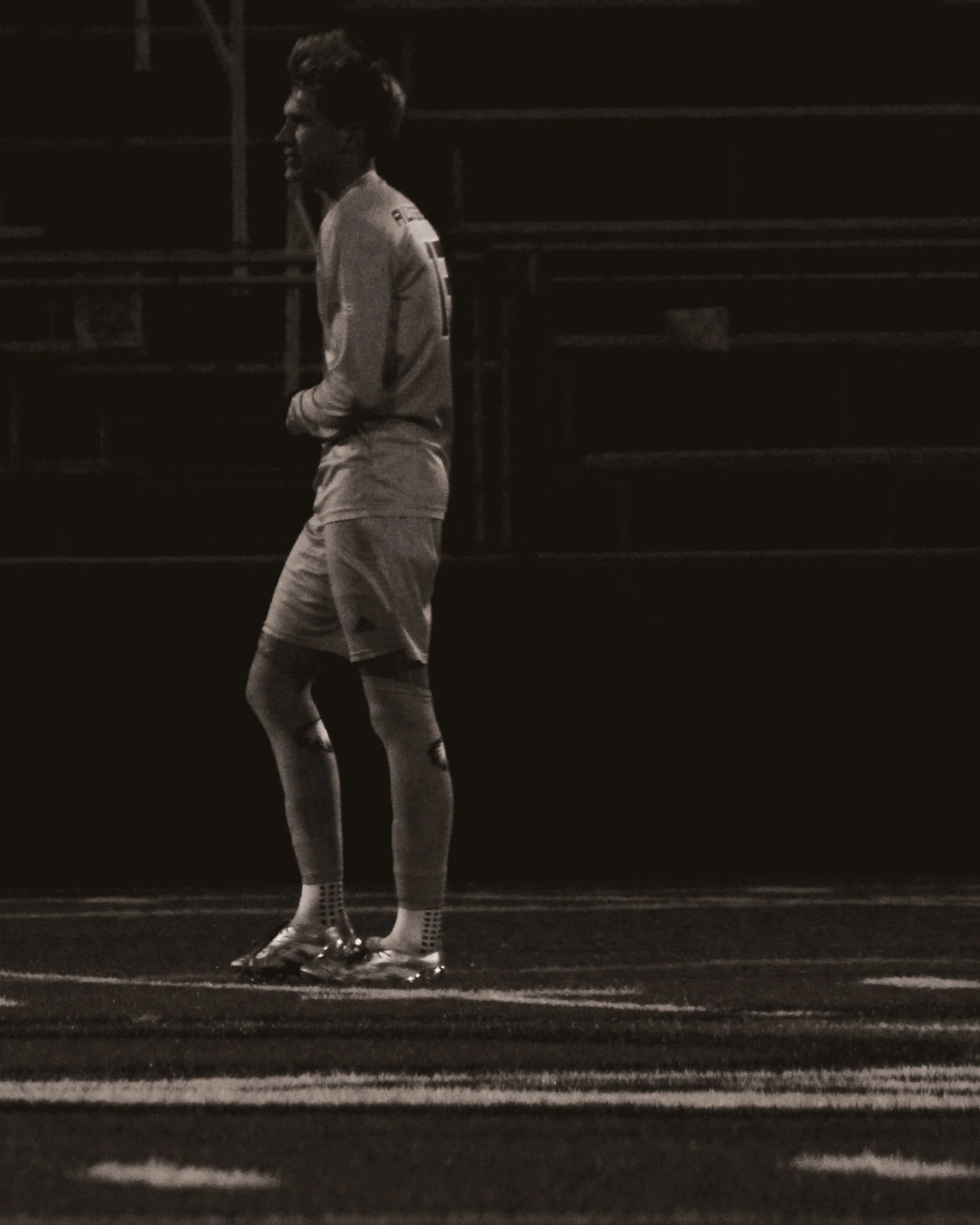A male soccer player standing alone on a field at night, wearing a long-sleeve jersey, shorts, and cleats.