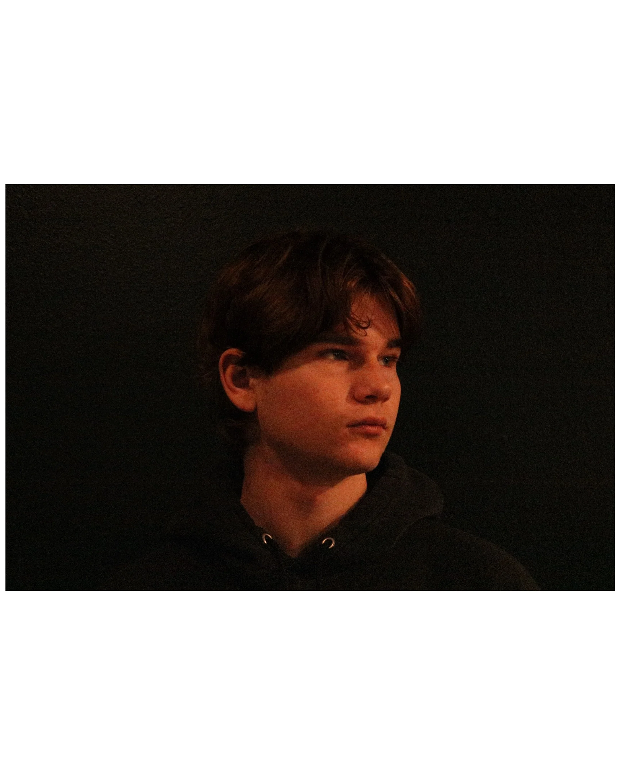 A young man with brown hair and fair skin looking to the side against a dark background.