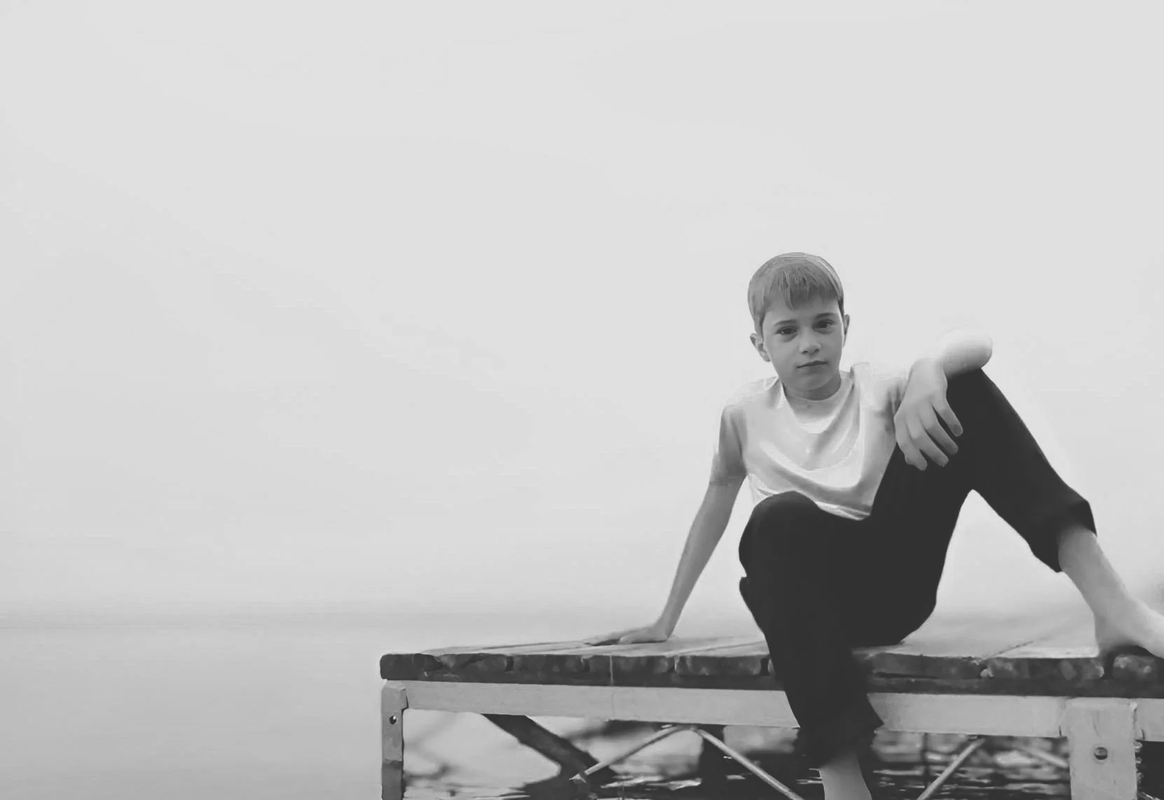 A boy sitting on the edge of a small dock, one foot in the water, looking at the camera, in black and white.