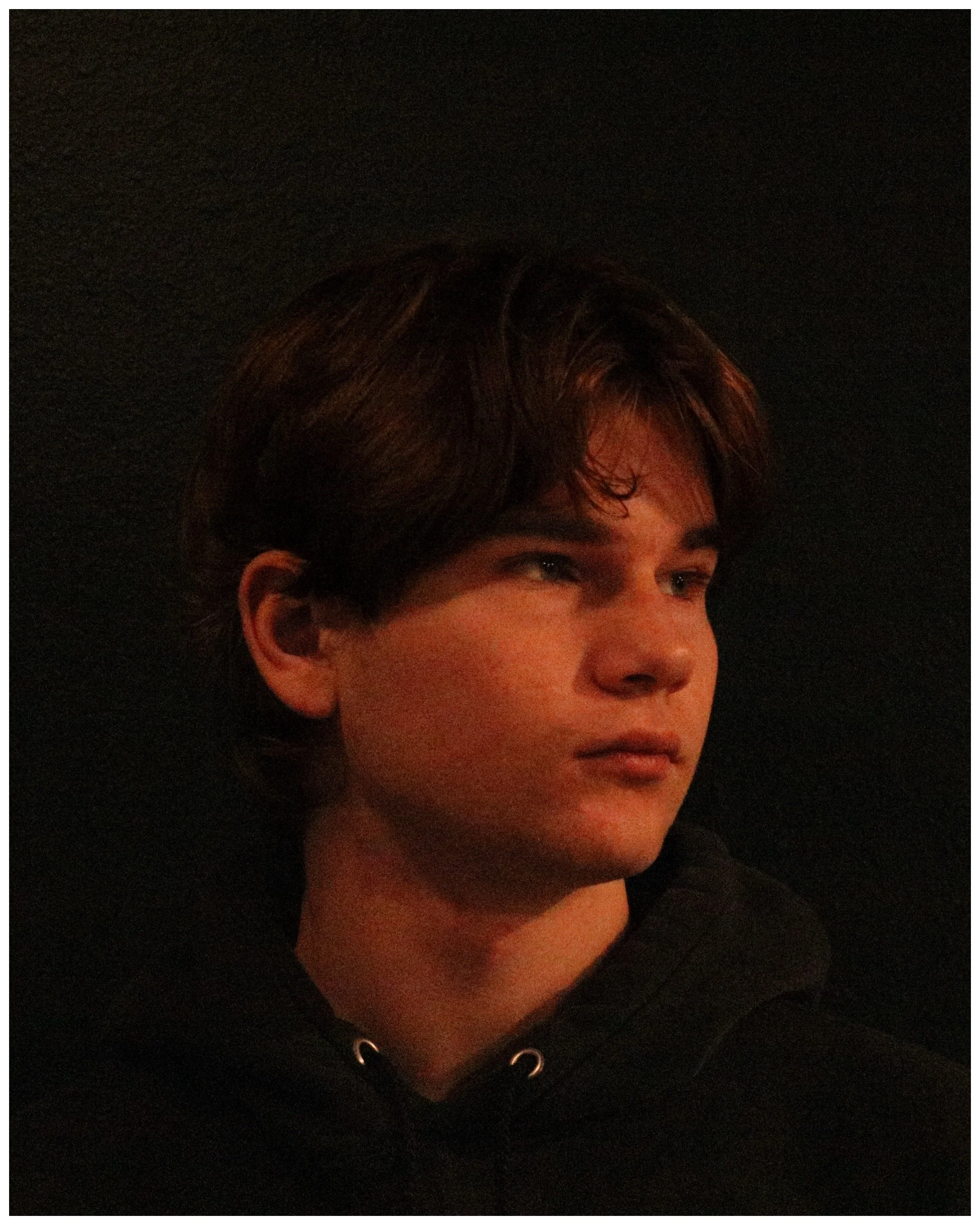 A young man with brown hair looking to the right, wearing a black hoodie, against a dark background.