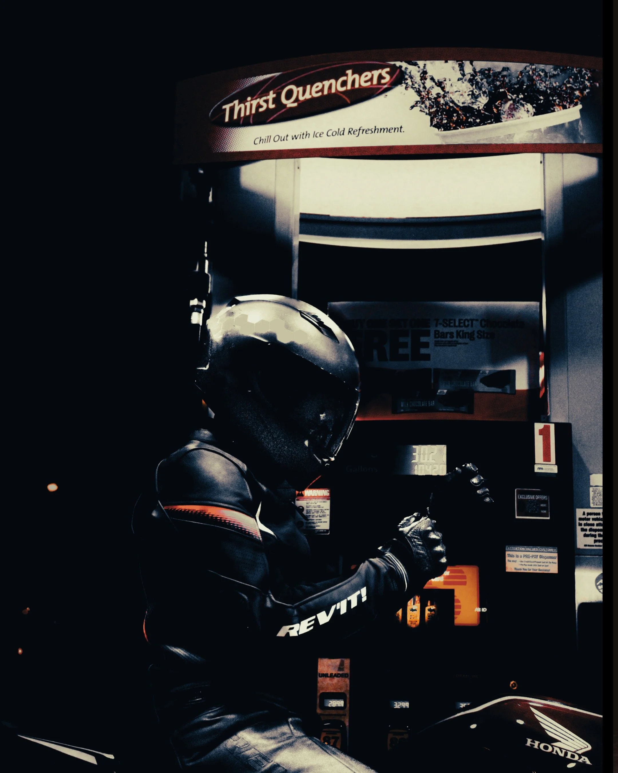 A motorcyclist wearing a black helmet and leather jacket with Revit branding, sitting by a gas pump at night. The gas station sign reads 'Thirst Quenchers' and offers ice cold refreshments.