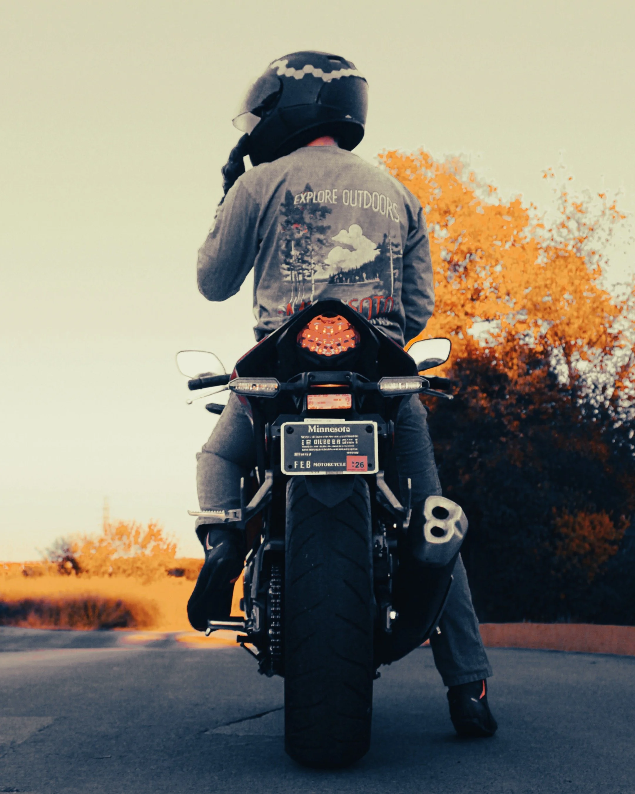 A person wearing a helmet and motorcycle gear standing on a black motorcycle on an open road with orange autumn trees in the background.