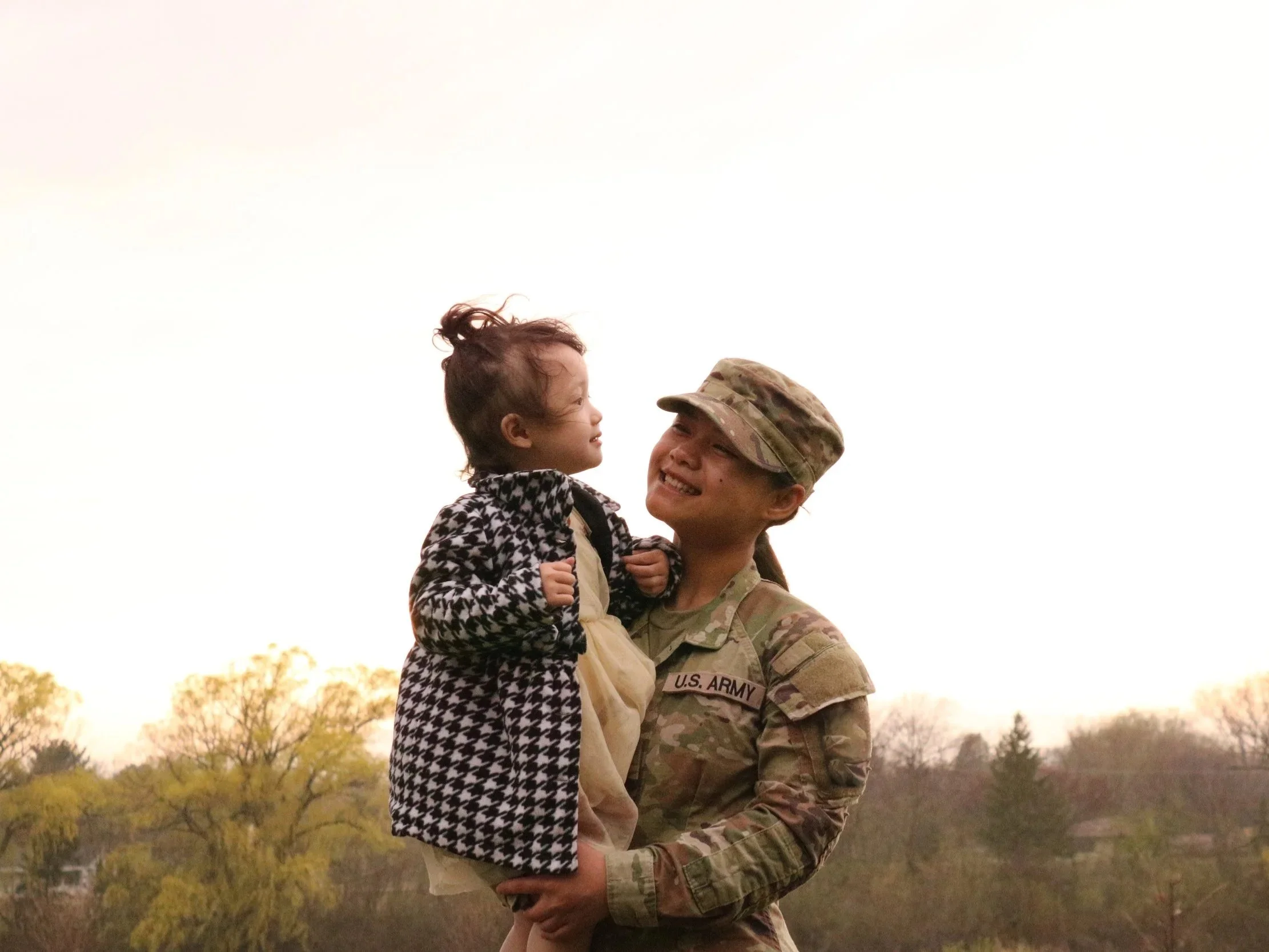 A U.S. Army soldier holding a young girl outdoors during sunset, smiling and looking at each other.