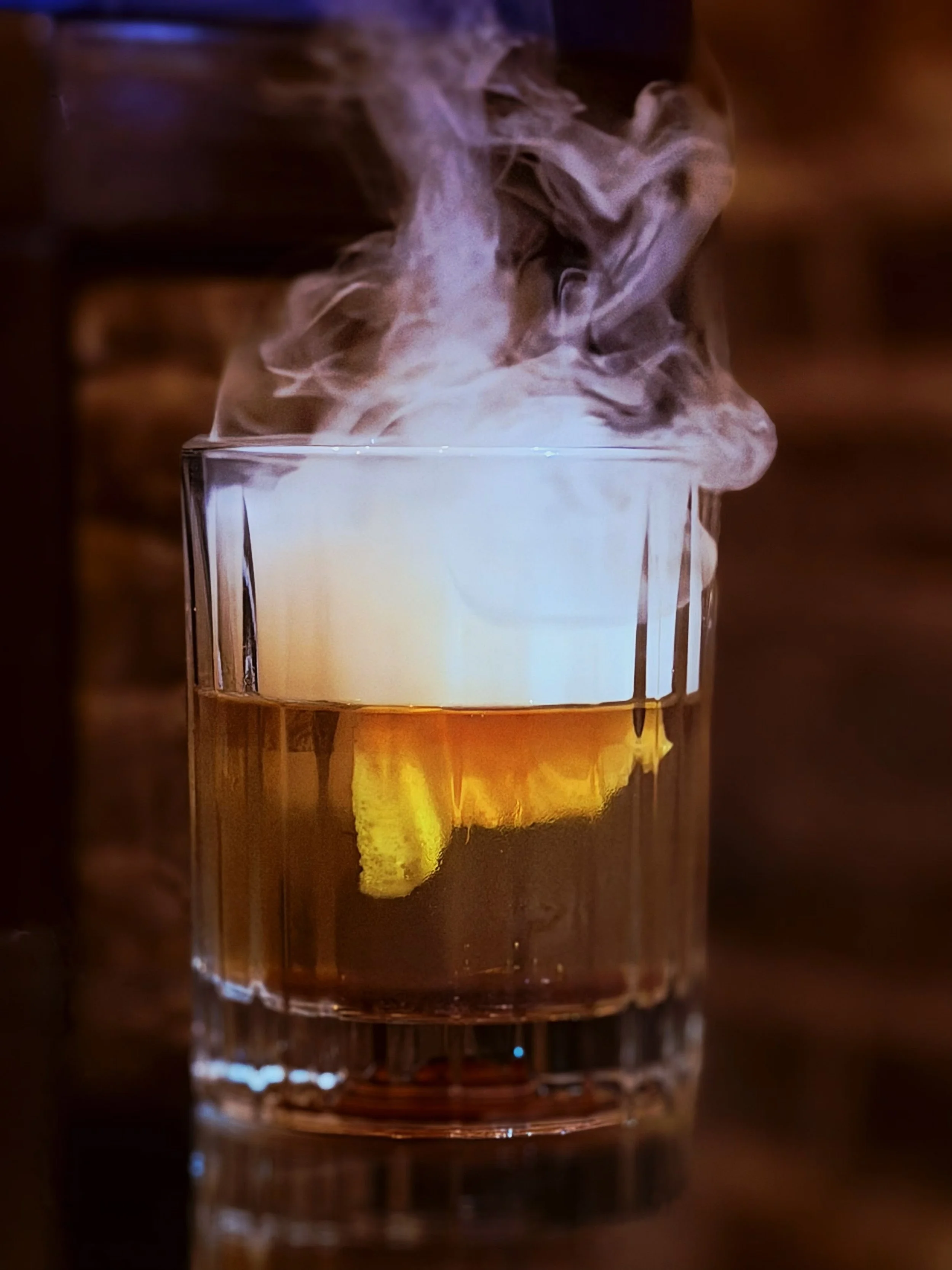 A glass of whiskey with a lemon wedge, with smoke or vapor rising from it, against a blurred dark background.