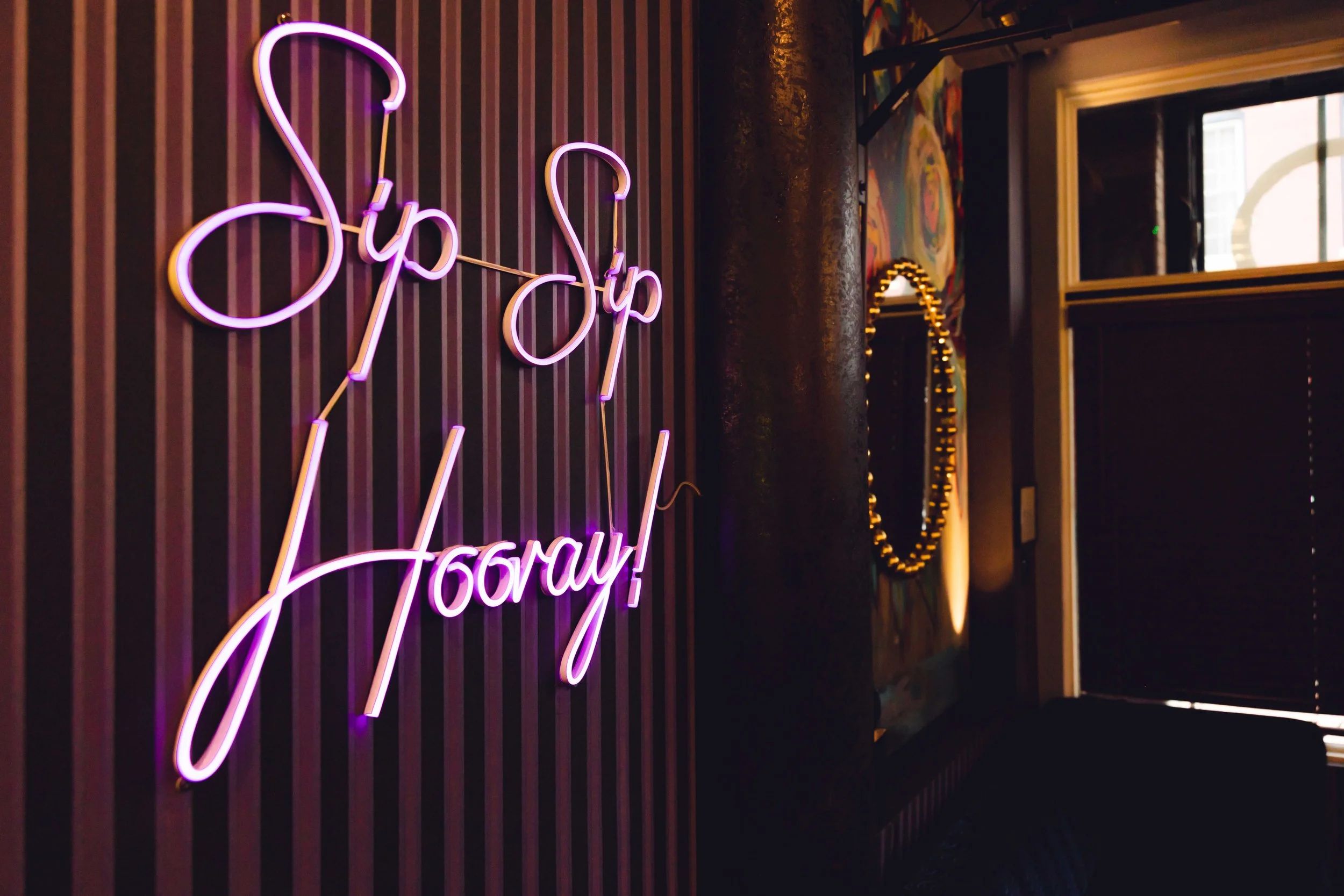 Neon sign on a wall that reads 'Sip Sip Hooray!' in pink and purple lights.