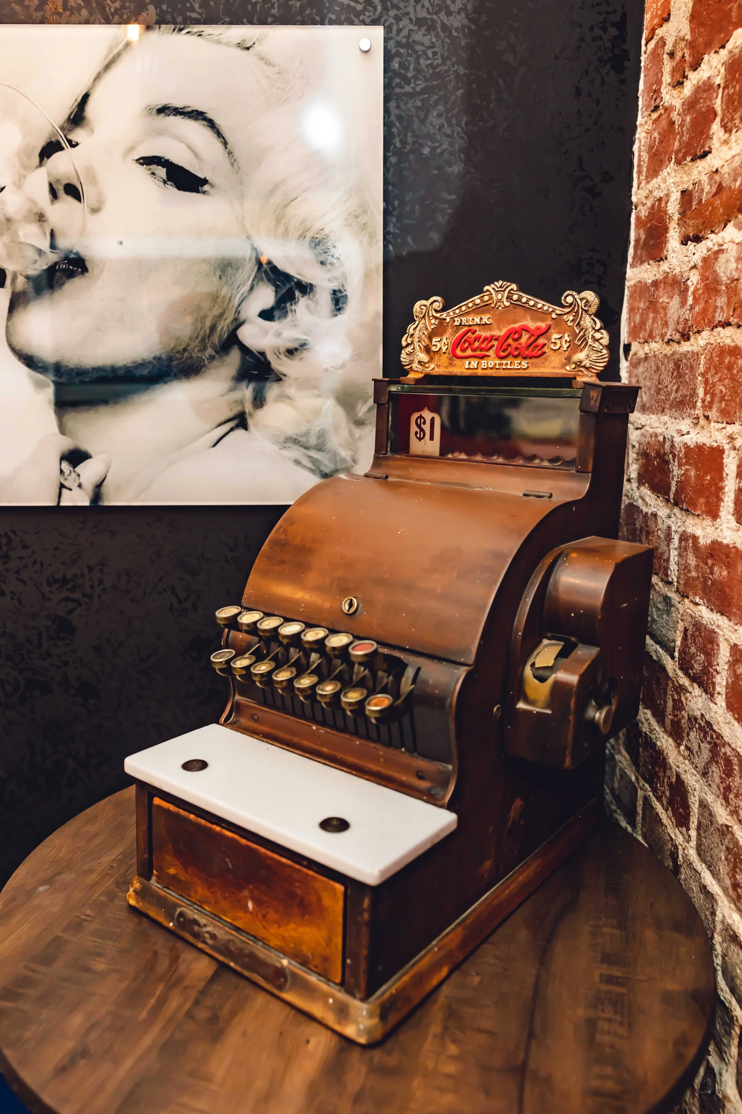 Vintage cash register with a Coca-Cola sign on top, display showing one dollar, and a black and white portrait of Marilyn Monroe on the wall behind it.
