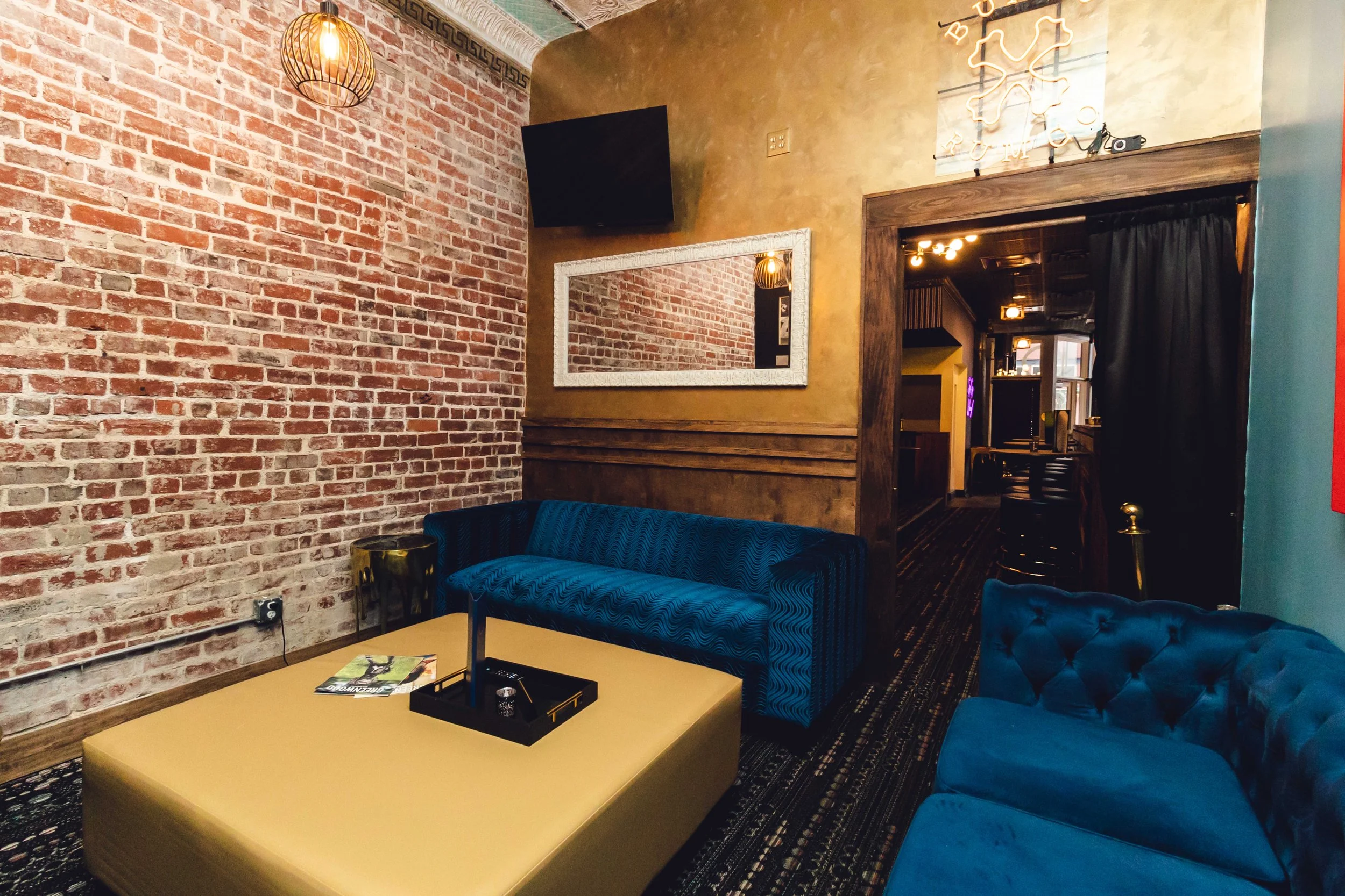 Interior of a cozy lounge area with a brick wall, a blue velvet sofa, a yellow ottoman with a magazine and black decorative item on it, a wall mirror, and warm lighting.