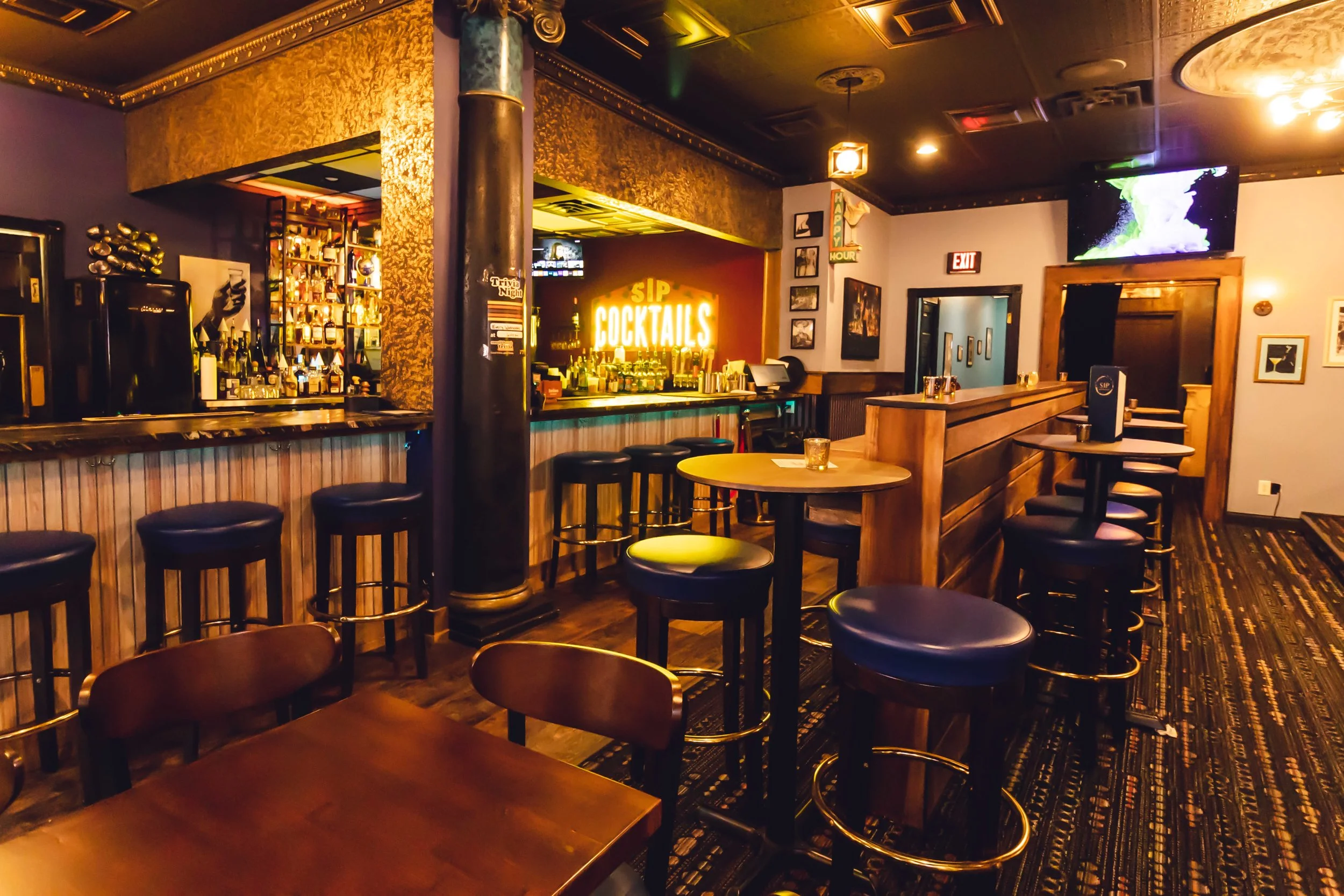 Interior of a dimly lit bar featuring a wooden bar counter with stools, a sign that reads 'Sip Cocktails,' a television on the wall, and framed pictures.
