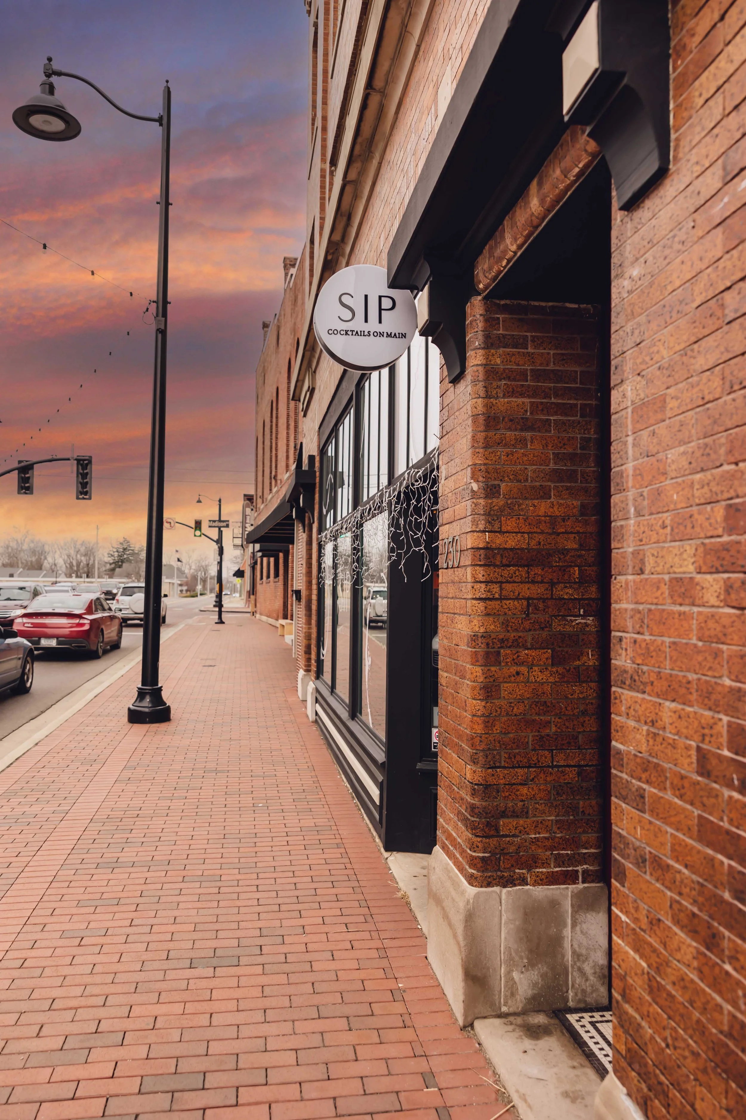 Street view of a brick building with a bar or restaurant named 'SIP' that serves cocktails, during sunset with a pink and orange sky, parked cars on the street, and street lamps.