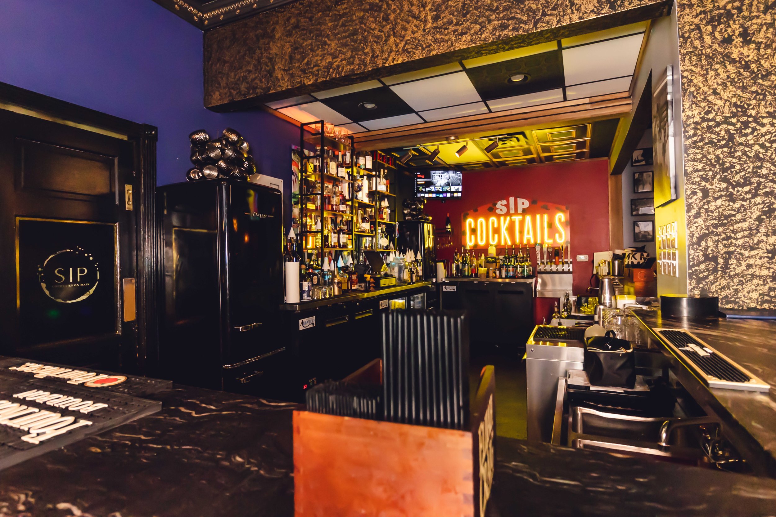 Inside a stylish bar with a neon sign reading 'SIP COCKTAILS' on a red wall. The bar counter has various bottles and bar equipment, with a backlit shelving unit displaying liquor bottles. The decor includes dark walls, textured gold patterns, and framed photographs.
