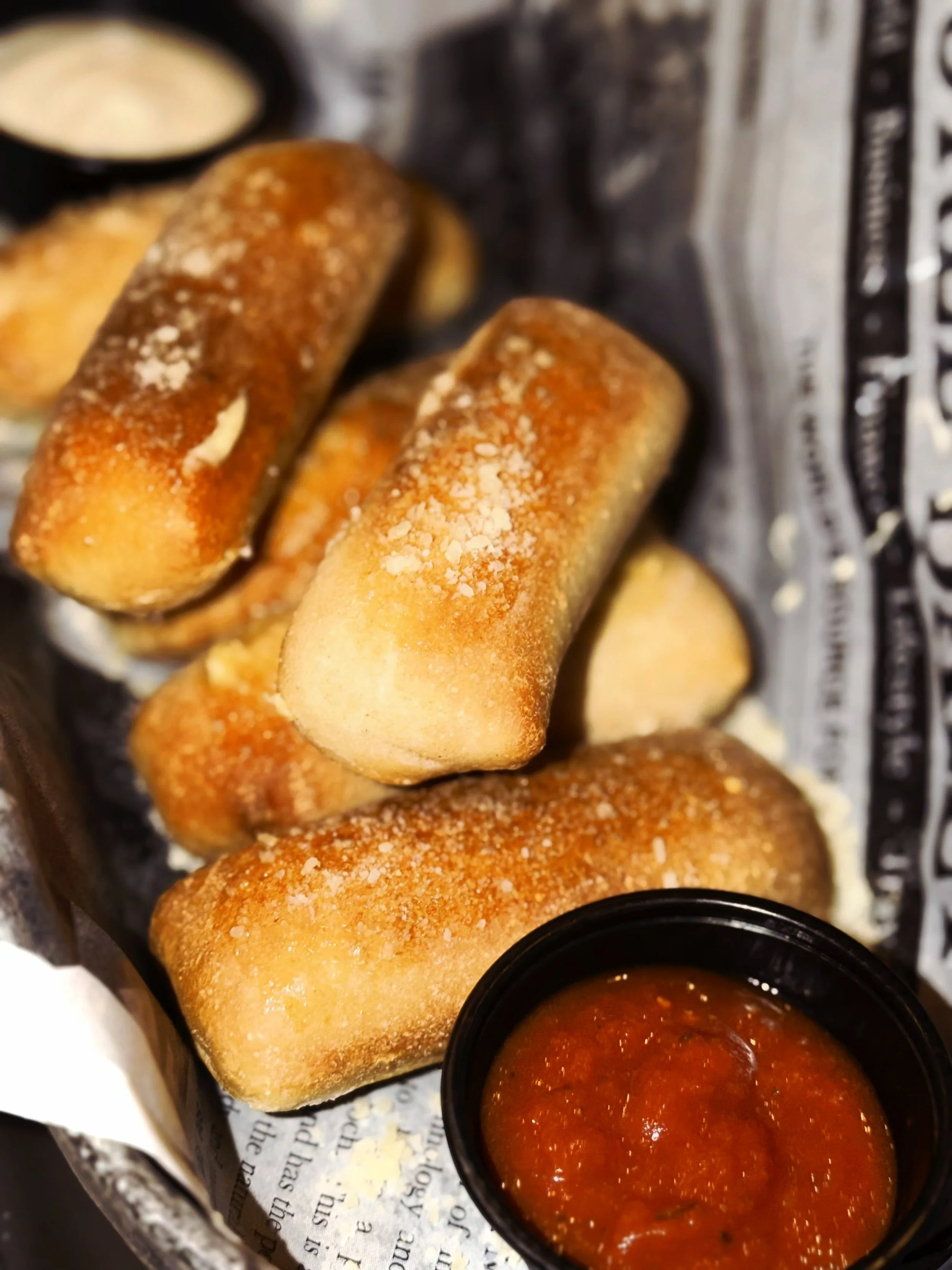 Four breadsticks with salt on top and a cup of marinara dipping sauce in a black container.