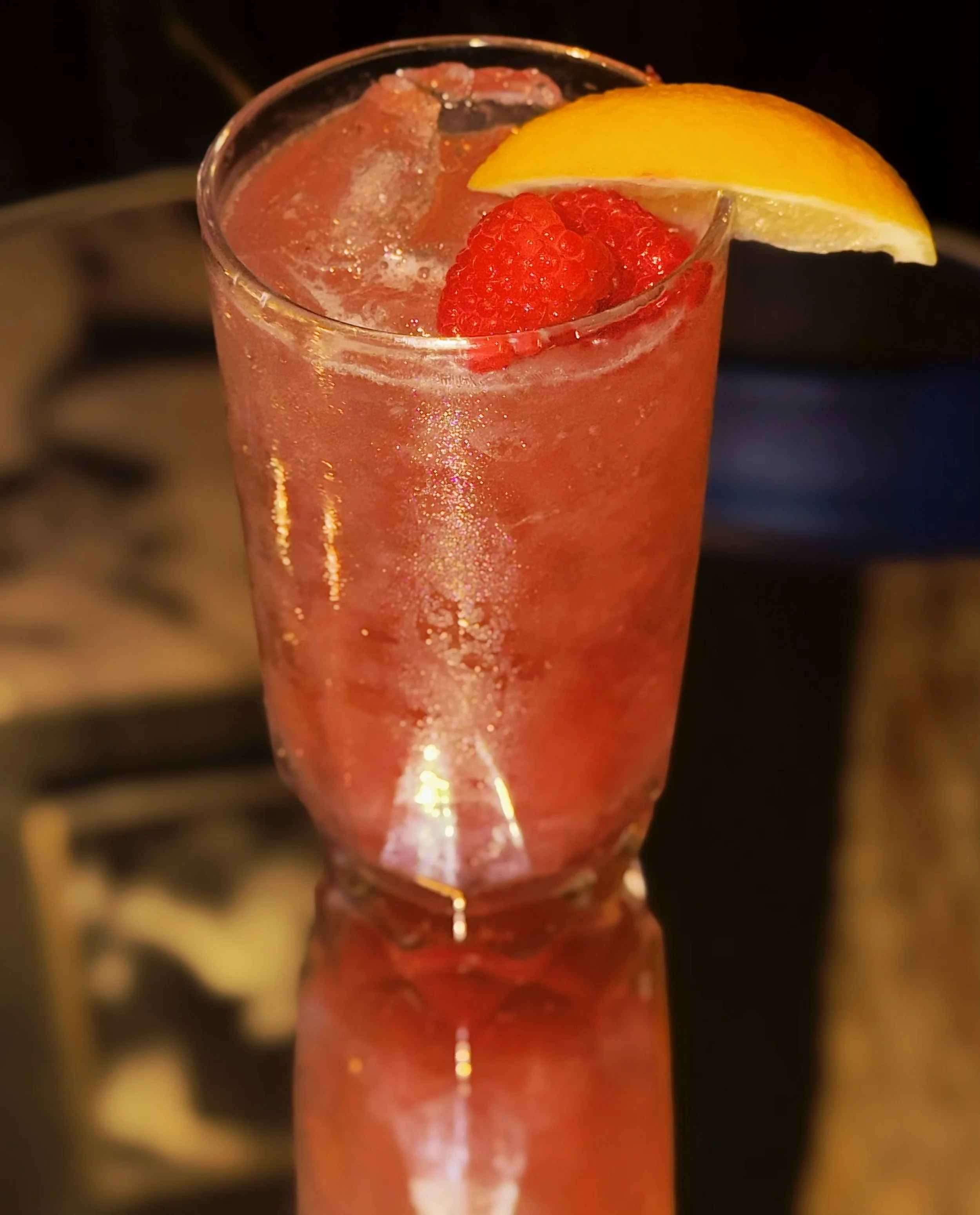 A tall glass of pink sparkling beverage garnished with a lemon wedge and red raspberries.