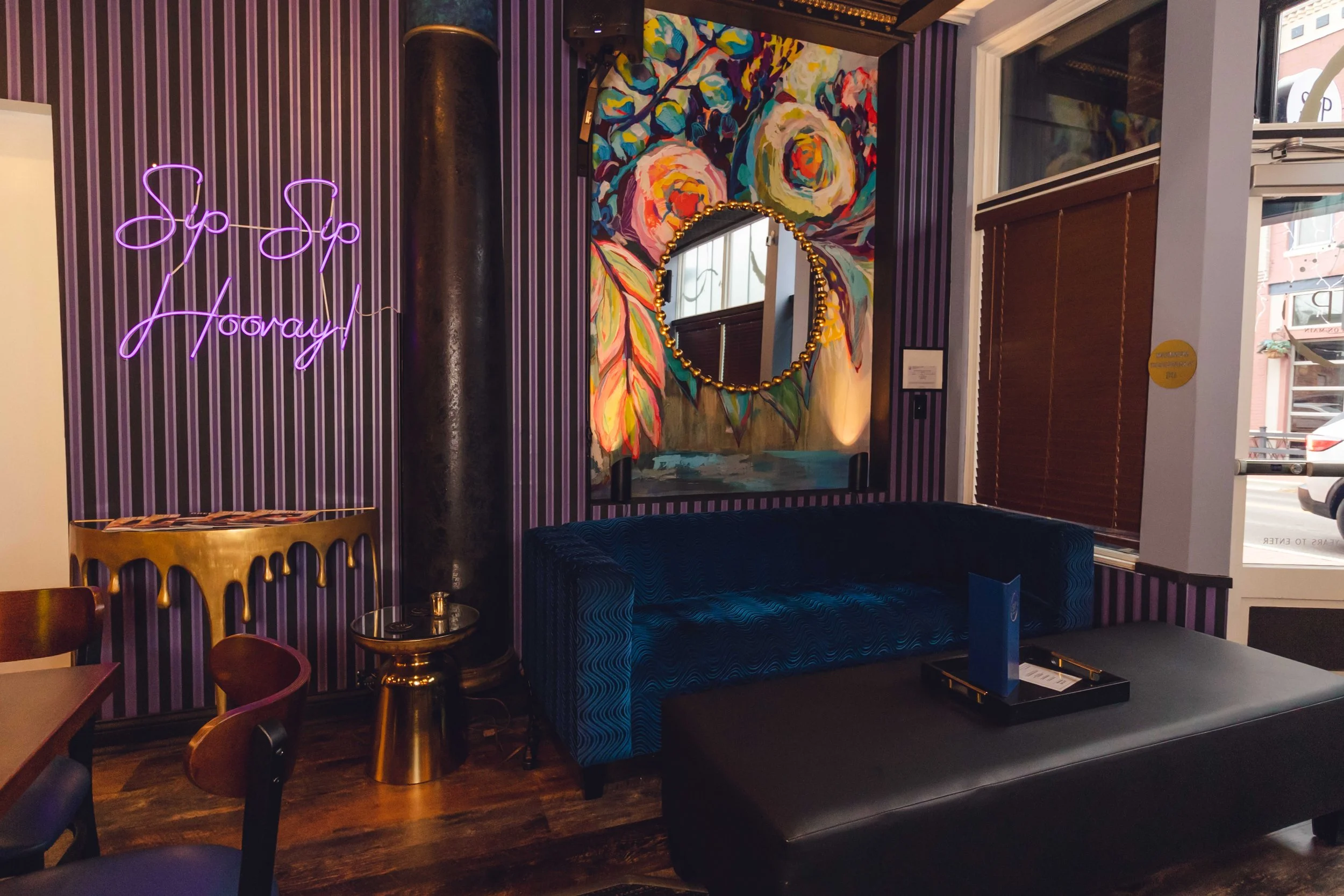 Interior of a cafe or lounge with purple-striped walls, neon purple sign saying 'Sip Sip Hooray,' a colorful abstract floral painting with a round mirror decorated with gold beads, a blue velvet sofa, a black ottoman with a menu, and wooden chairs and tables near a window with brown blinds.