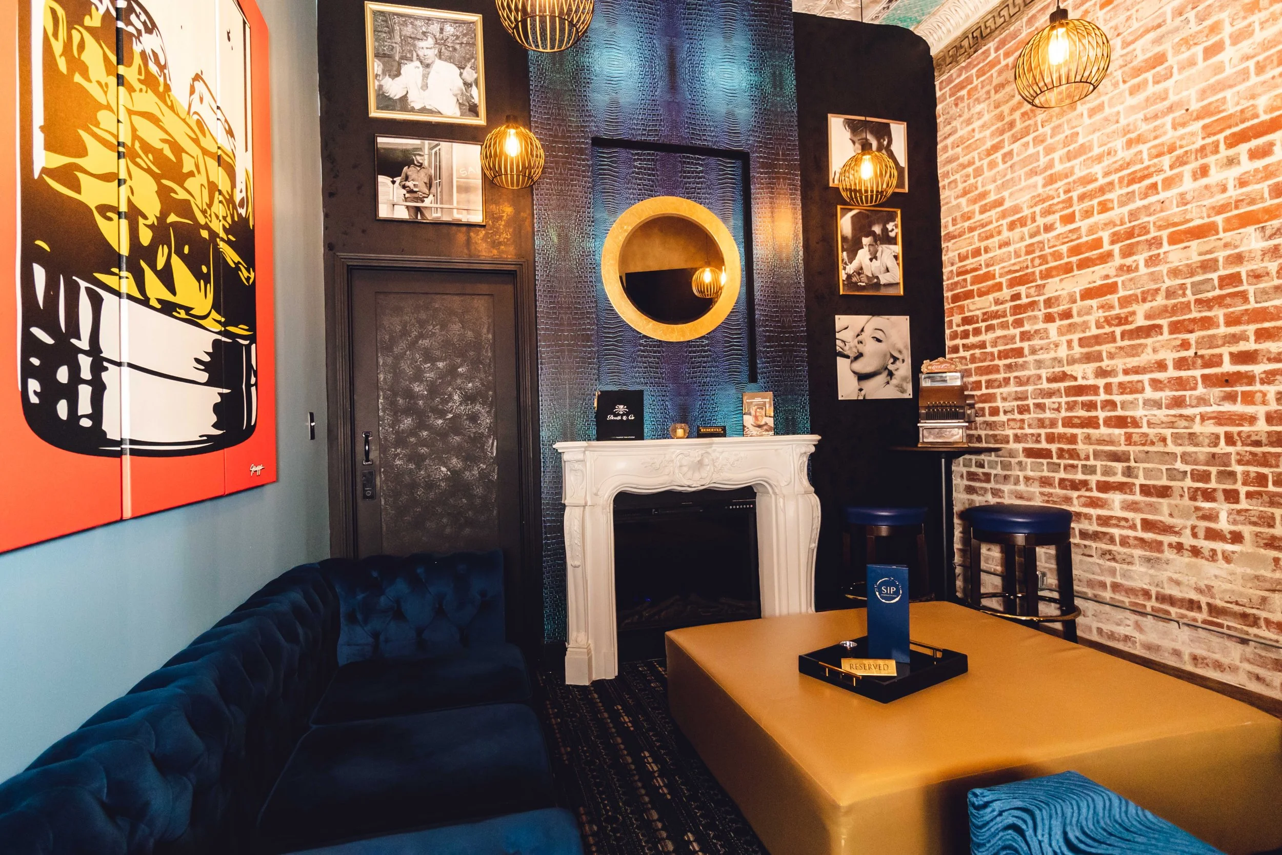 A modern lounge with a mix of vintage and contemporary decor. Features include a white fireplace, blue velvet sofa, a bright yellow ottoman, industrial hanging lights, black and white photographs, colorful artwork, and a red brick wall.