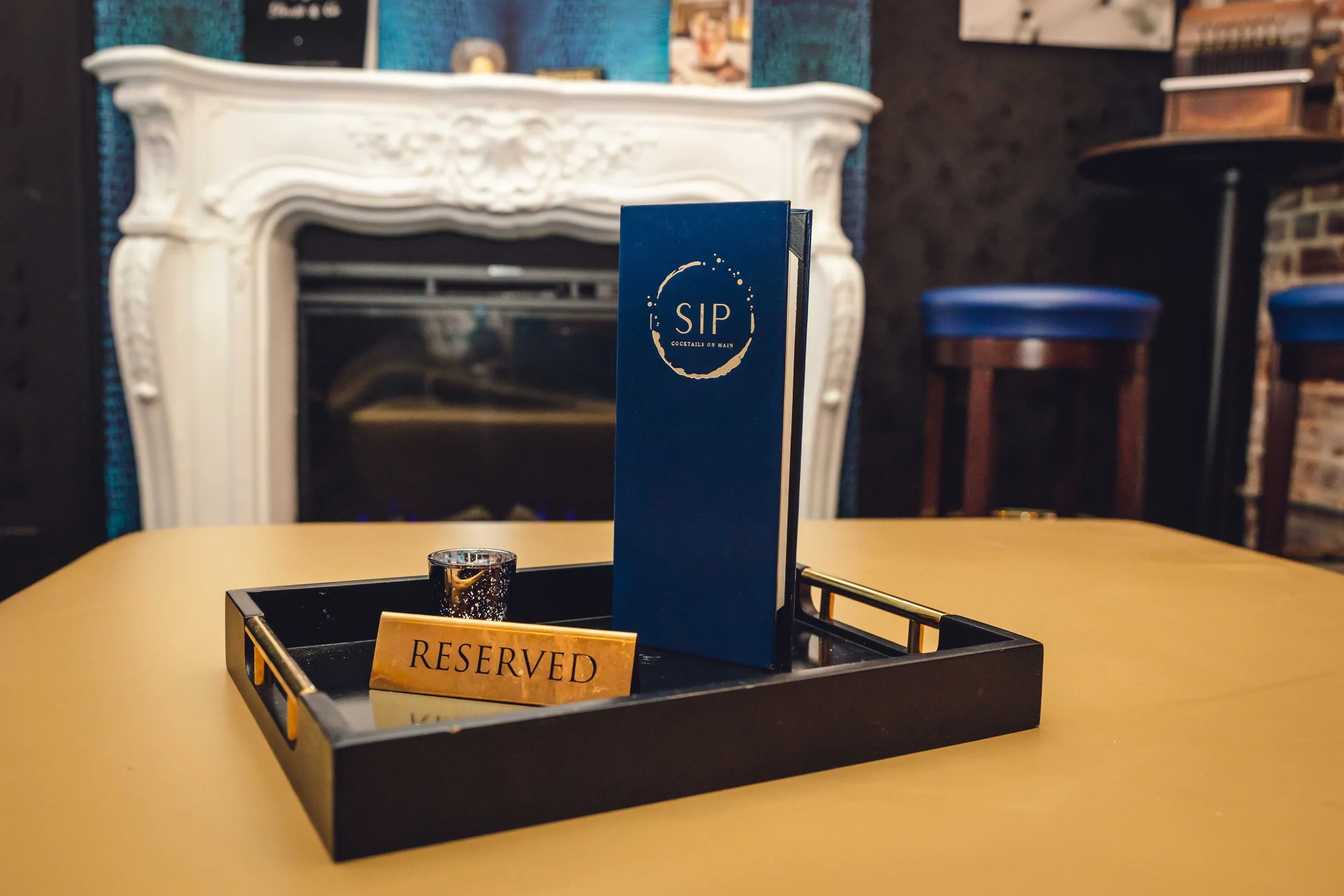 A table in a restaurant or bar with a black tray holding a reserved sign, a small candle, and a blue drink menu with the logo 'SIP Cocktails on Main'. In the background, there is a white ornate fireplace, a dark wall, and a blue barstool.
