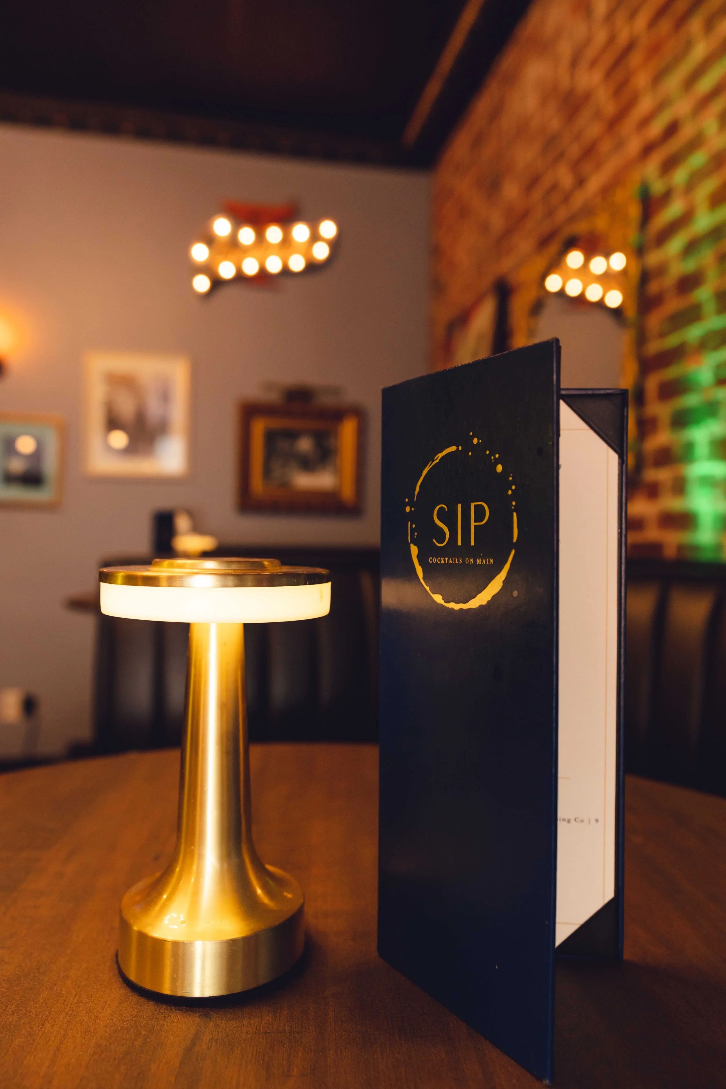 Close-up of a gold table lamp and a black menu with 'SIP Cocktails on Main' on a wooden table in a dimly lit restaurant or bar with framed pictures and wall lights in the background.