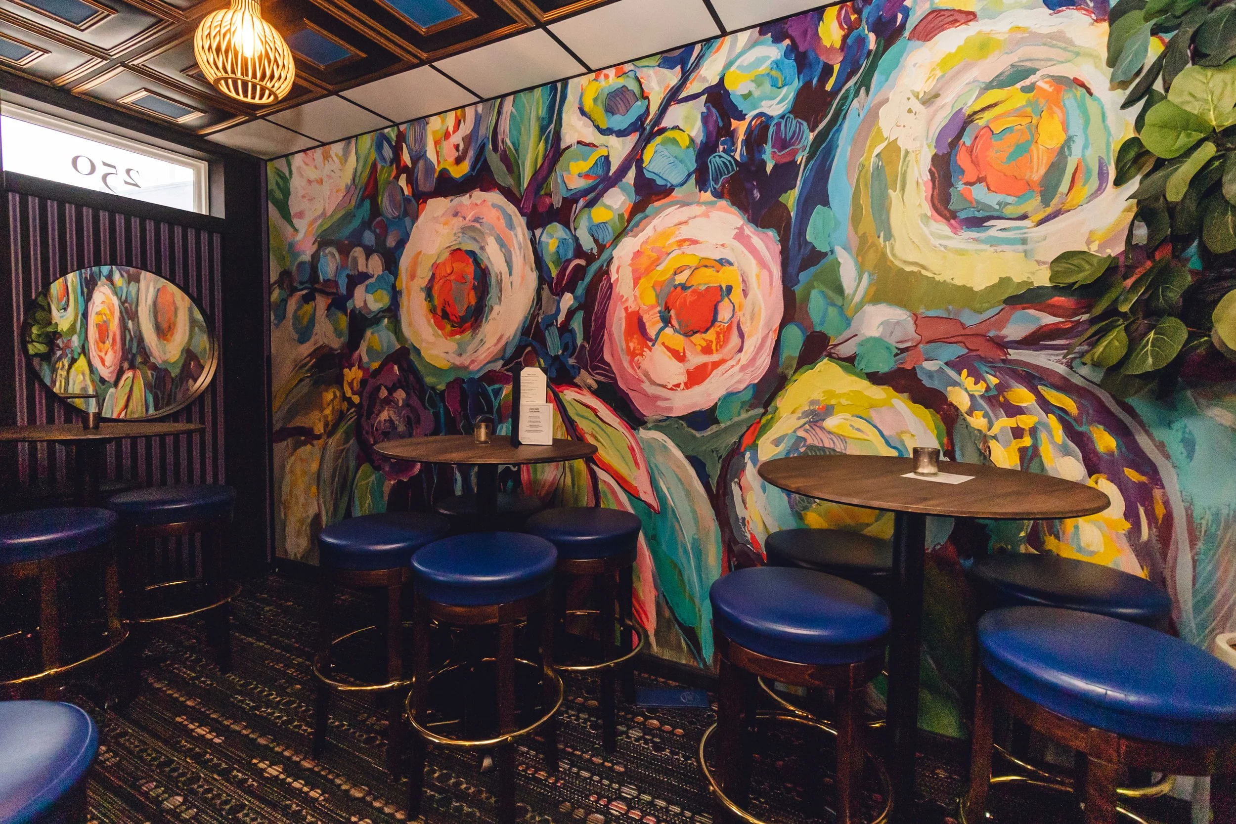 Interior of a restaurant with a large, colorful floral mural on the wall, round tables with dark wooden tops and blue cushioned stools, a decorative lamp hanging from the ceiling, a small mirror showing part of the mural, a dark patterned carpet, and a leafy green plant in the corner.