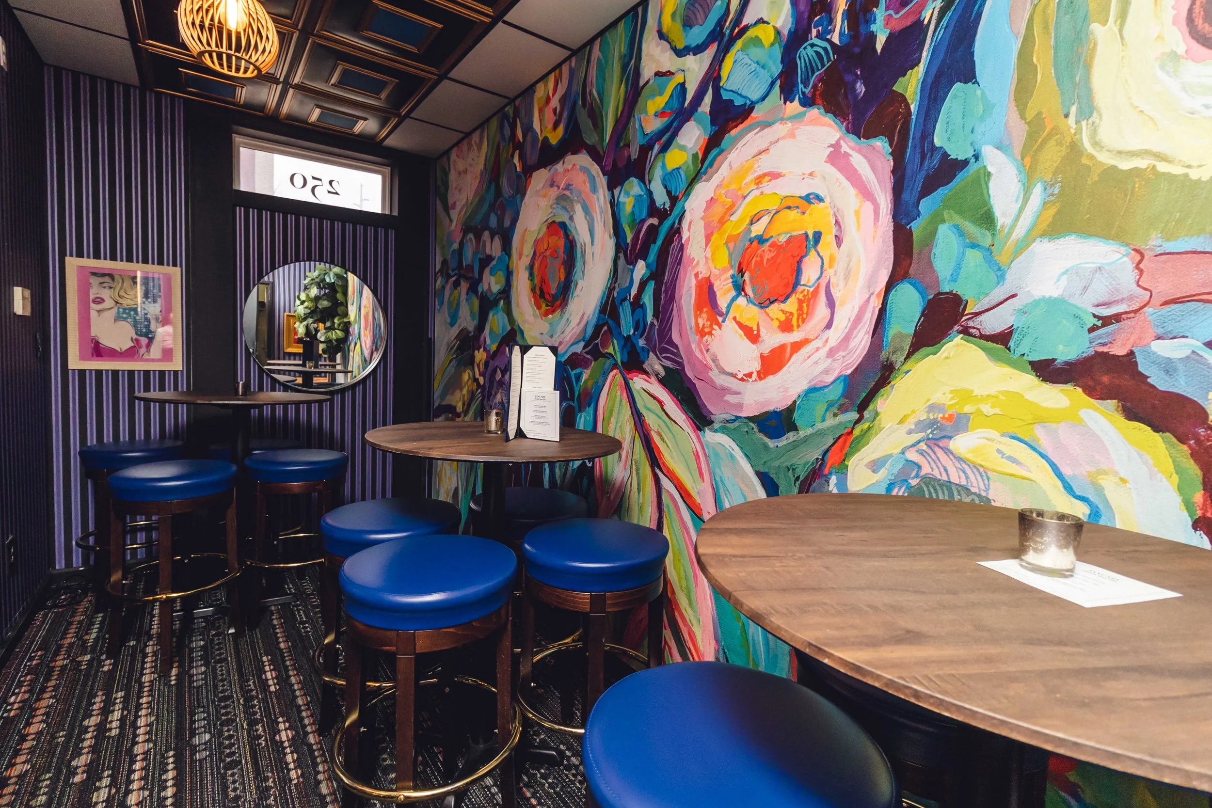 A corner of a restaurant with a vibrant floral wall mural, round wooden tables, and blue cushioned stools with gold legs. A decorative mirror, framed poster of a woman, and a window with a sign are visible.