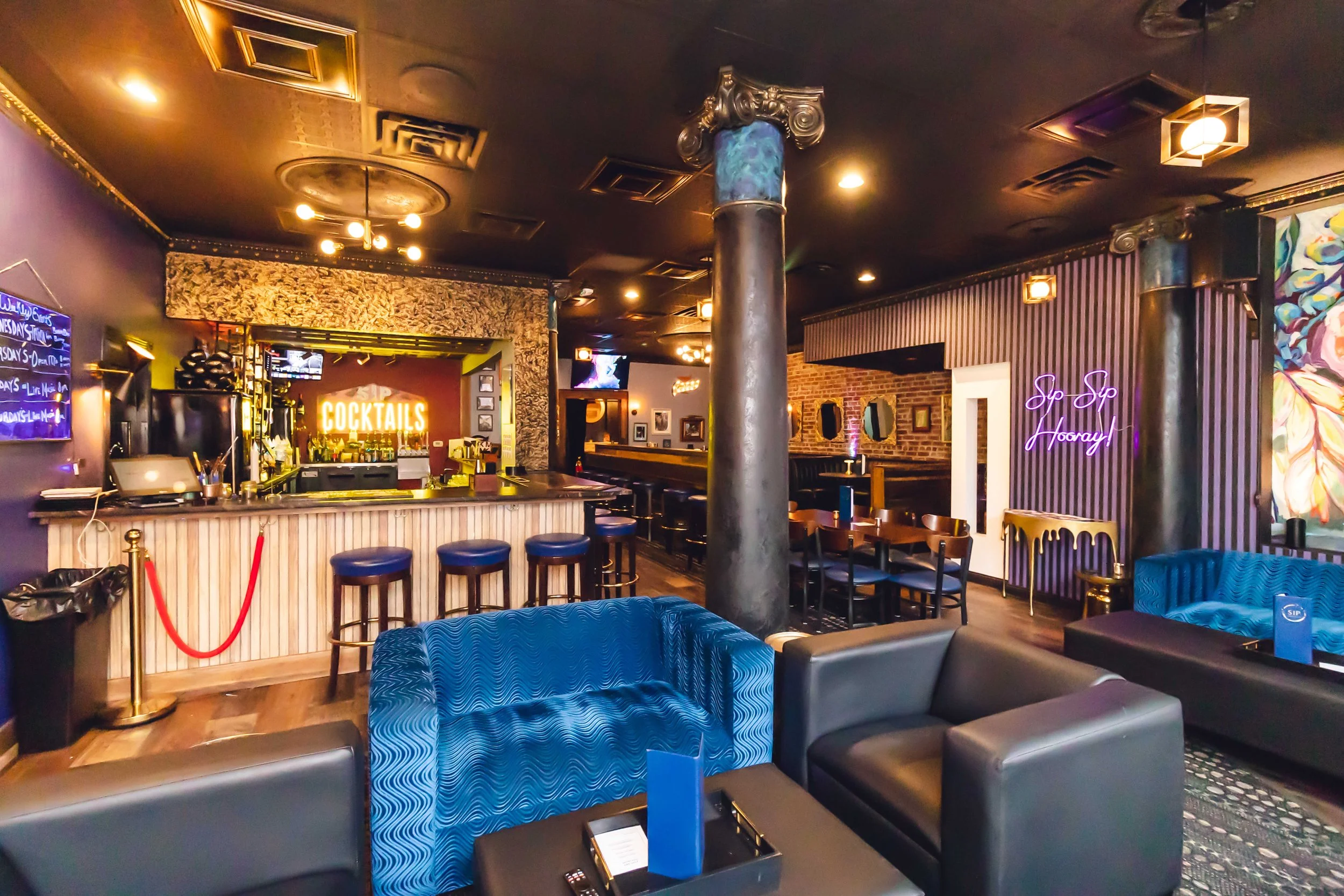 Interior of a bar with a purple and black color scheme, blue sofas, a counter with stools, neon signs that say 'COCKTAILS' and 'Sip Sip Hooray!'.