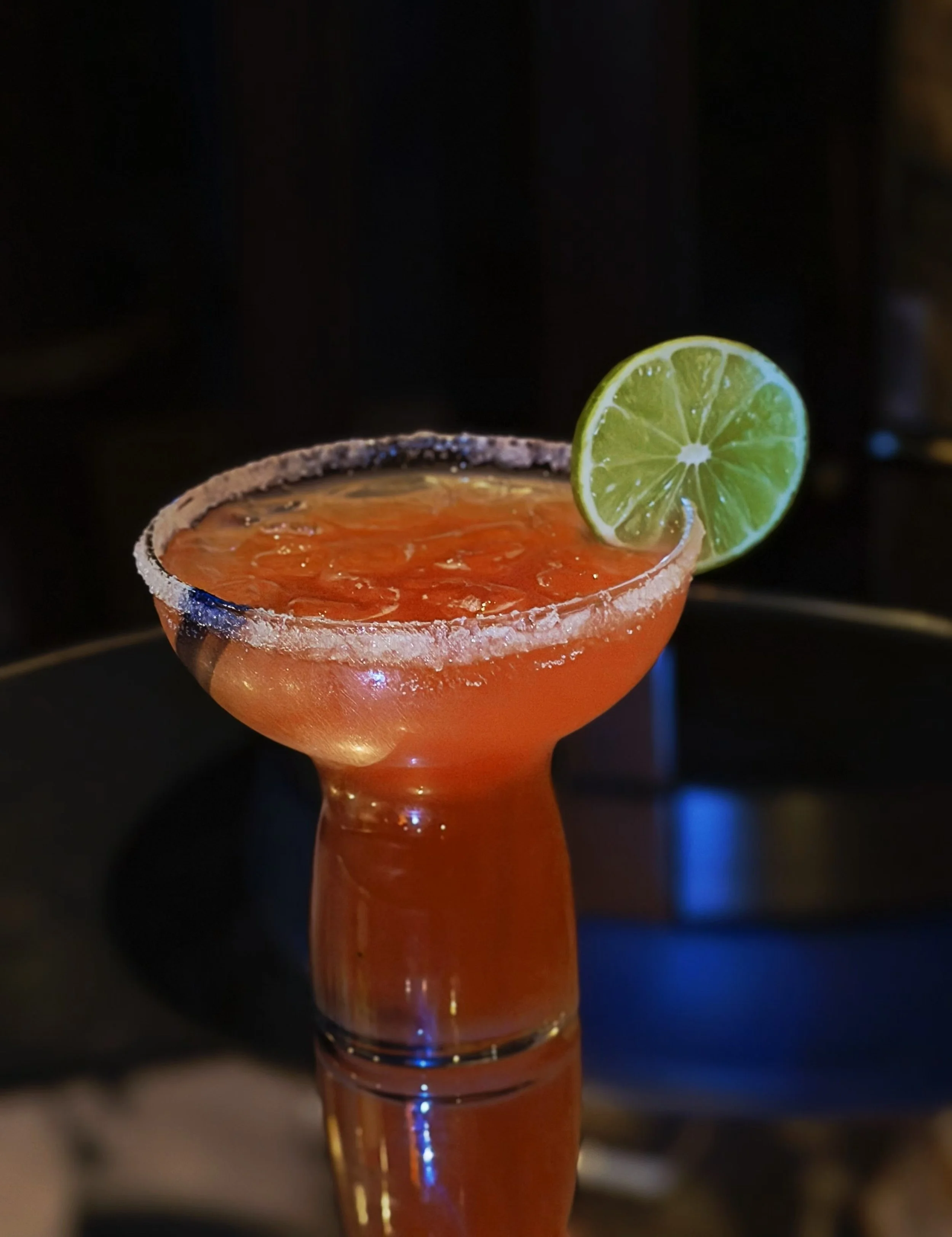 A cocktail drink in a glass with a salted rim, garnished with a lime slice on the rim, on a black surface with a dark background.