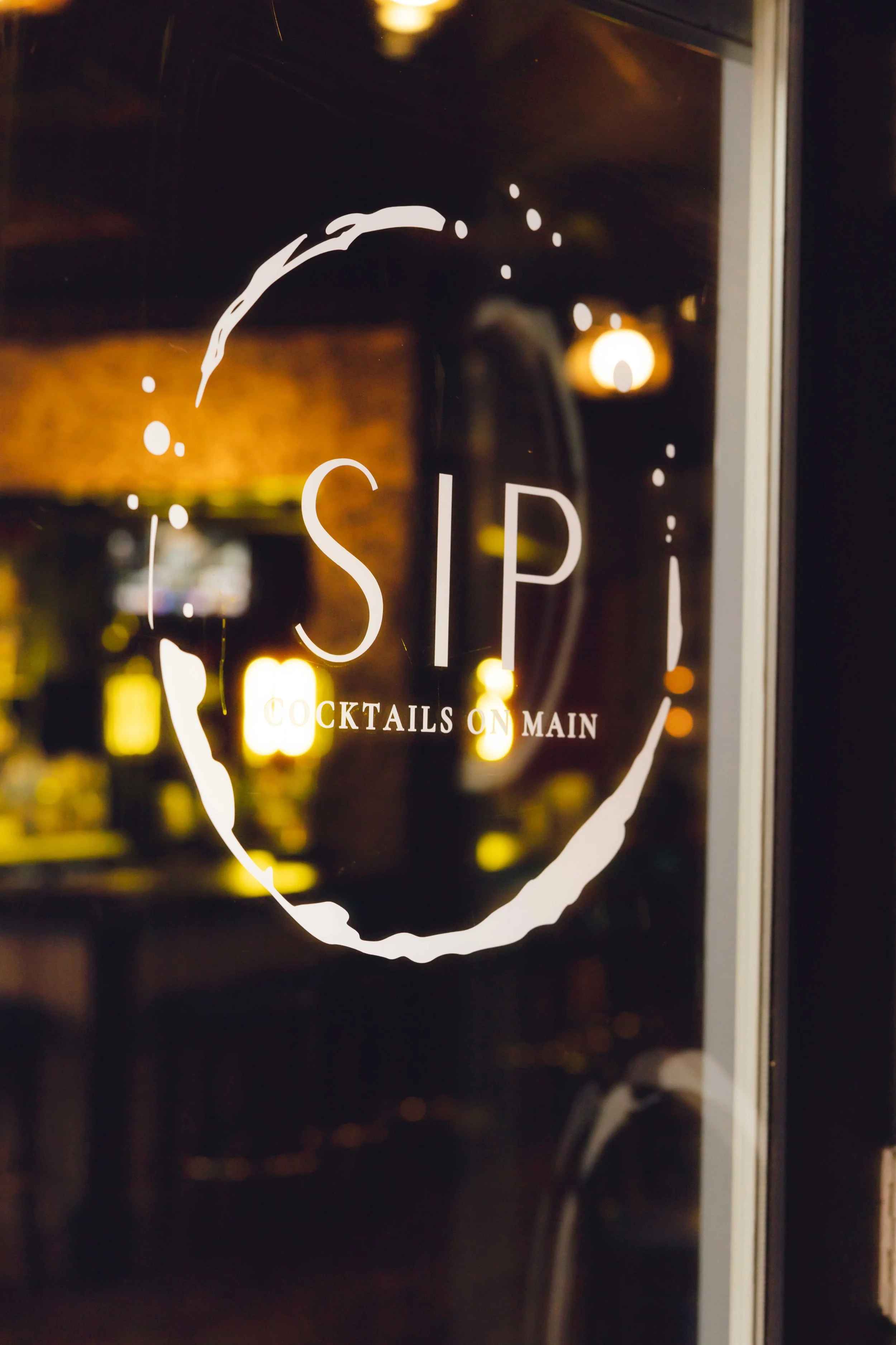 Restaurant window with the sign 'SIP' and the phrase 'Cocktails on Main' underneath, featuring a circular design around the text.