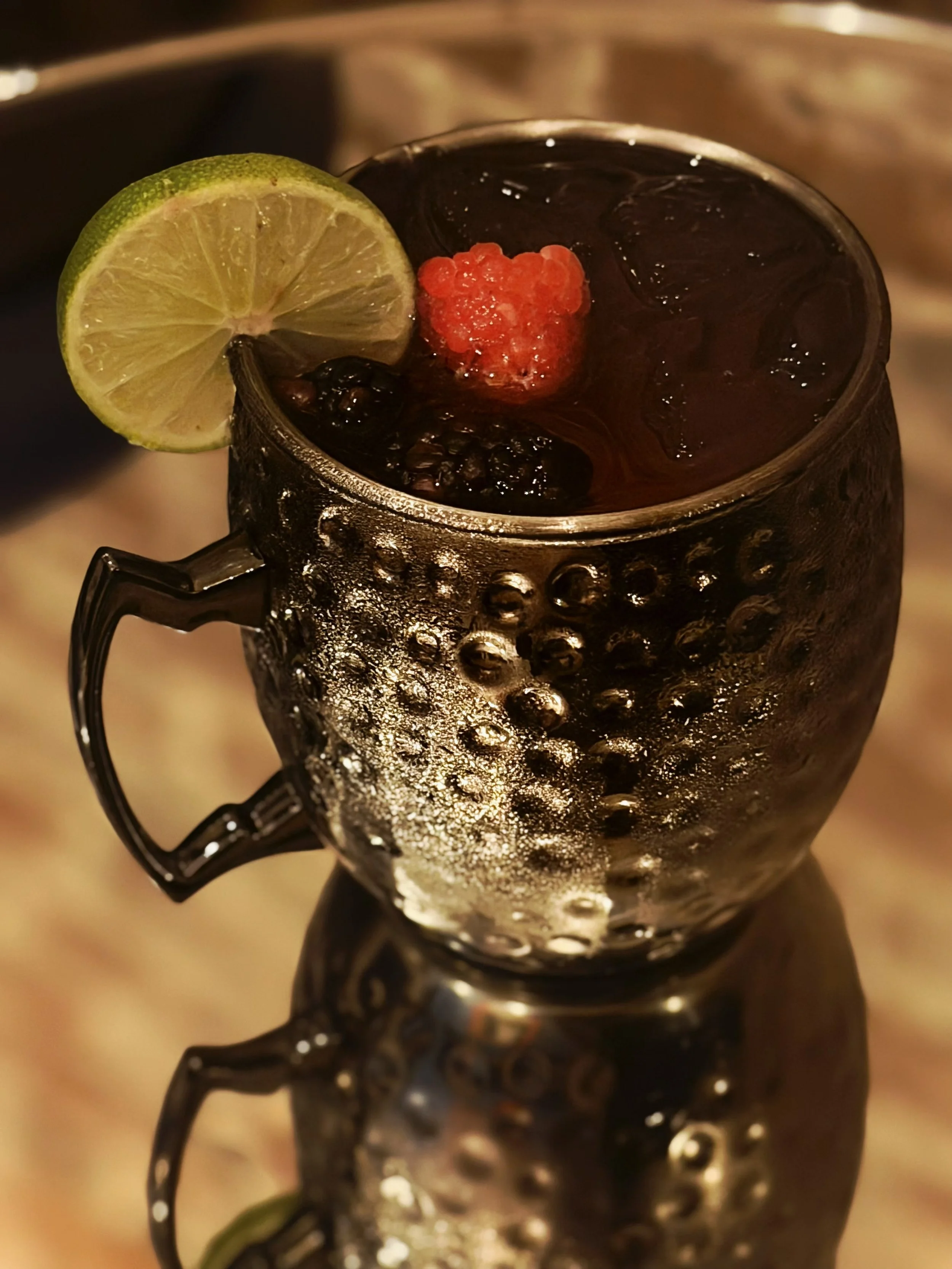 A metal cup filled with a dark beverage, ice, blackberries, and a red berry, garnished with a lime wedge on the rim, with a reflective surface and textured exterior.