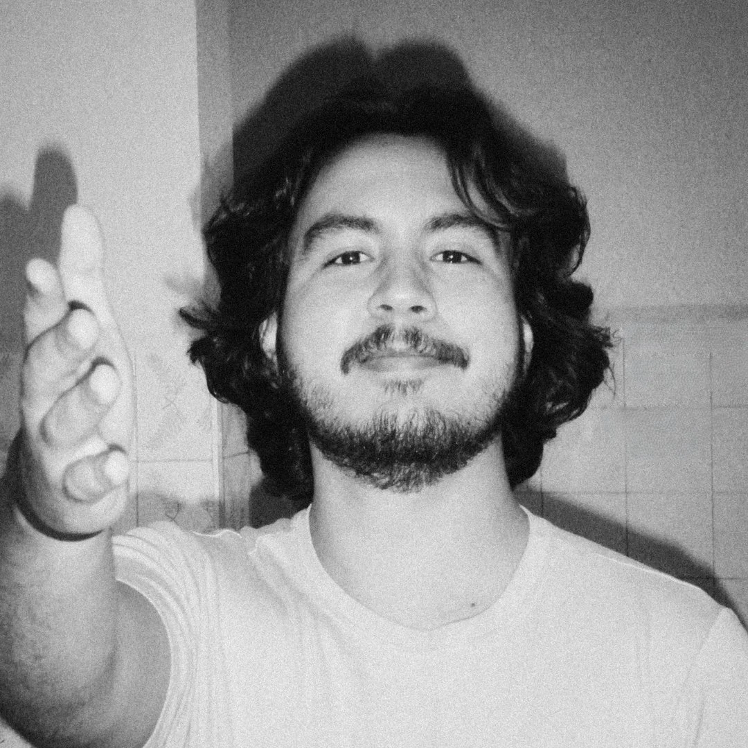 Homem de cabelo cacheado, barba e bigode, sorrindo e apontando para a câmera, em um ambiente interno
