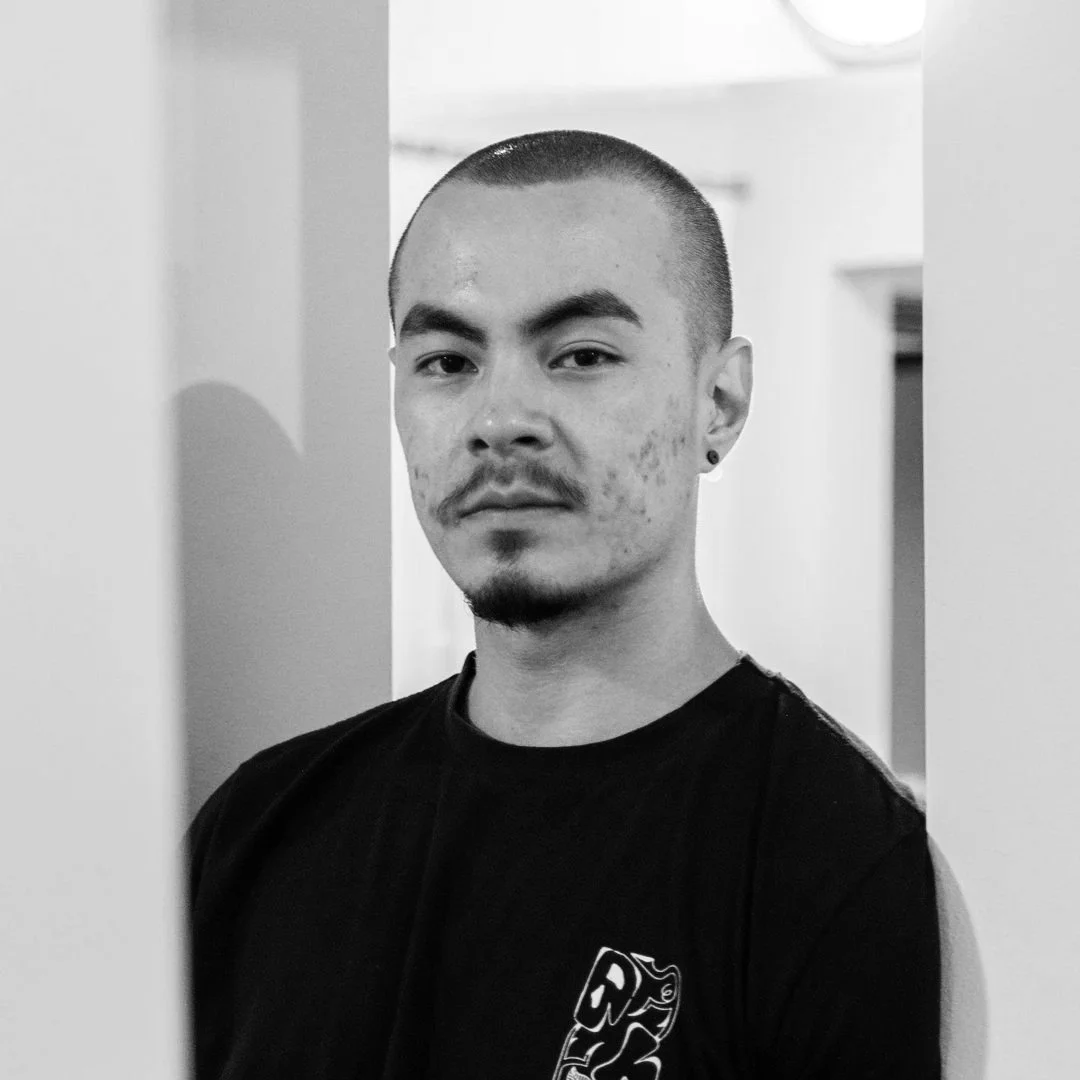 Retrato em preto e branco de um homem com cabelo raspado, barba e piercing na orelha, usando uma camiseta preta, em um ambiente interno.