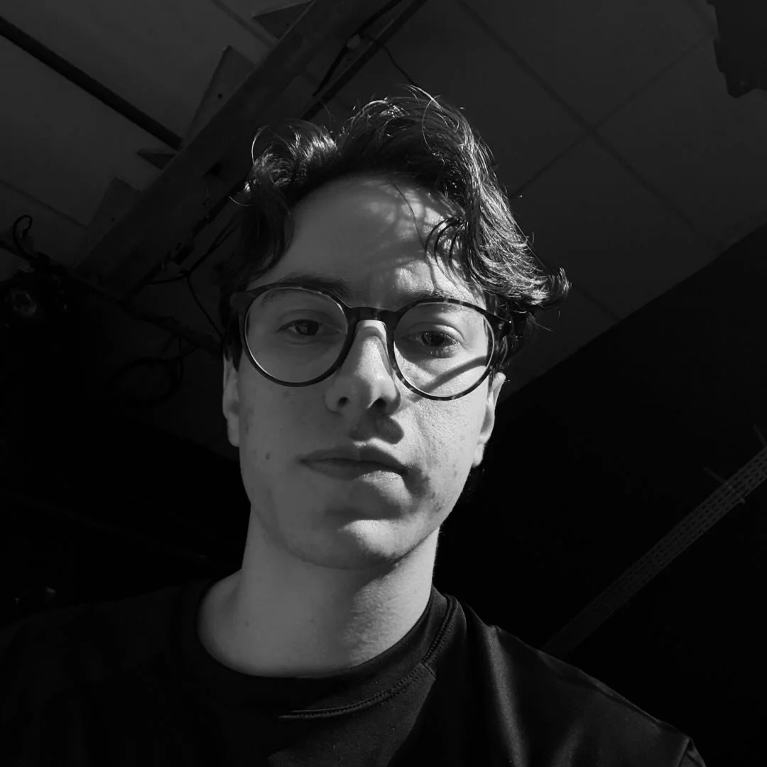 Retrato em preto e branco de uma pessoa com óculos grandes, cabelos ondulados e expressão neutra, com iluminação focada no rosto.