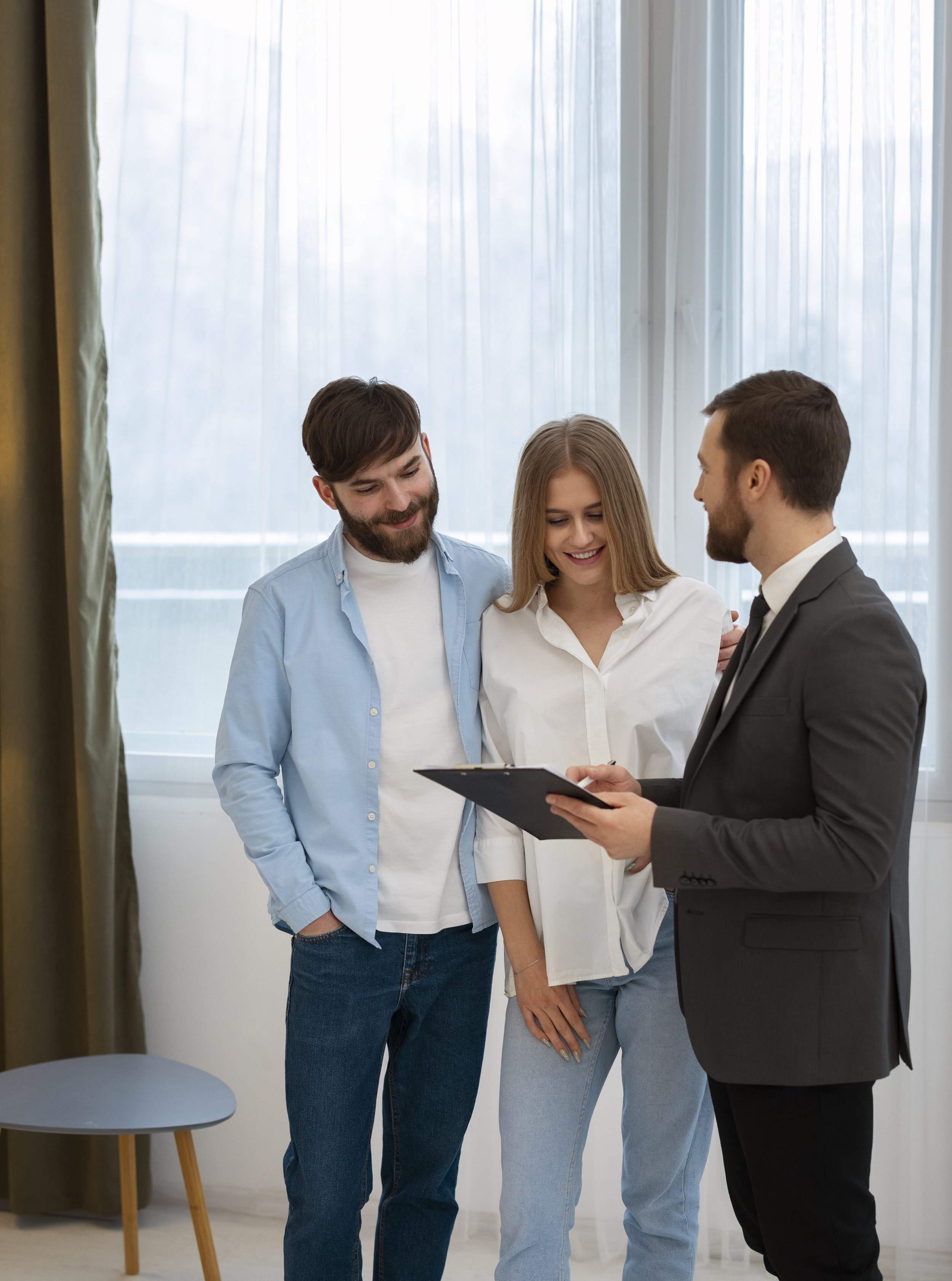 A real estate agent showing a house to a young couple, with all of them smiling and looking at a clipboard.