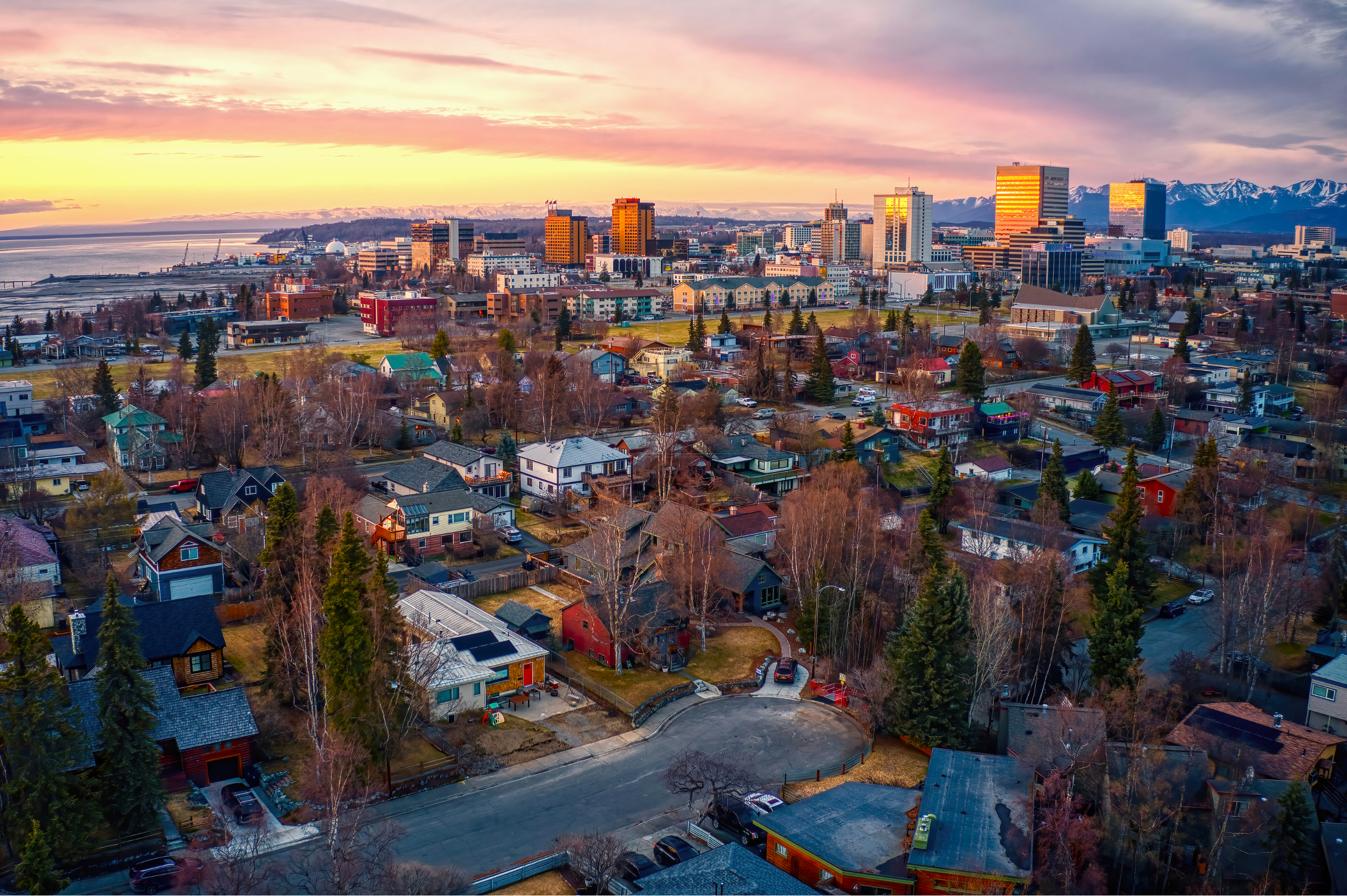 Aerial view of a city skyline at sunset with a mix of residential houses in the foreground and tall commercial buildings in the background, snow-capped mountains in the distance.