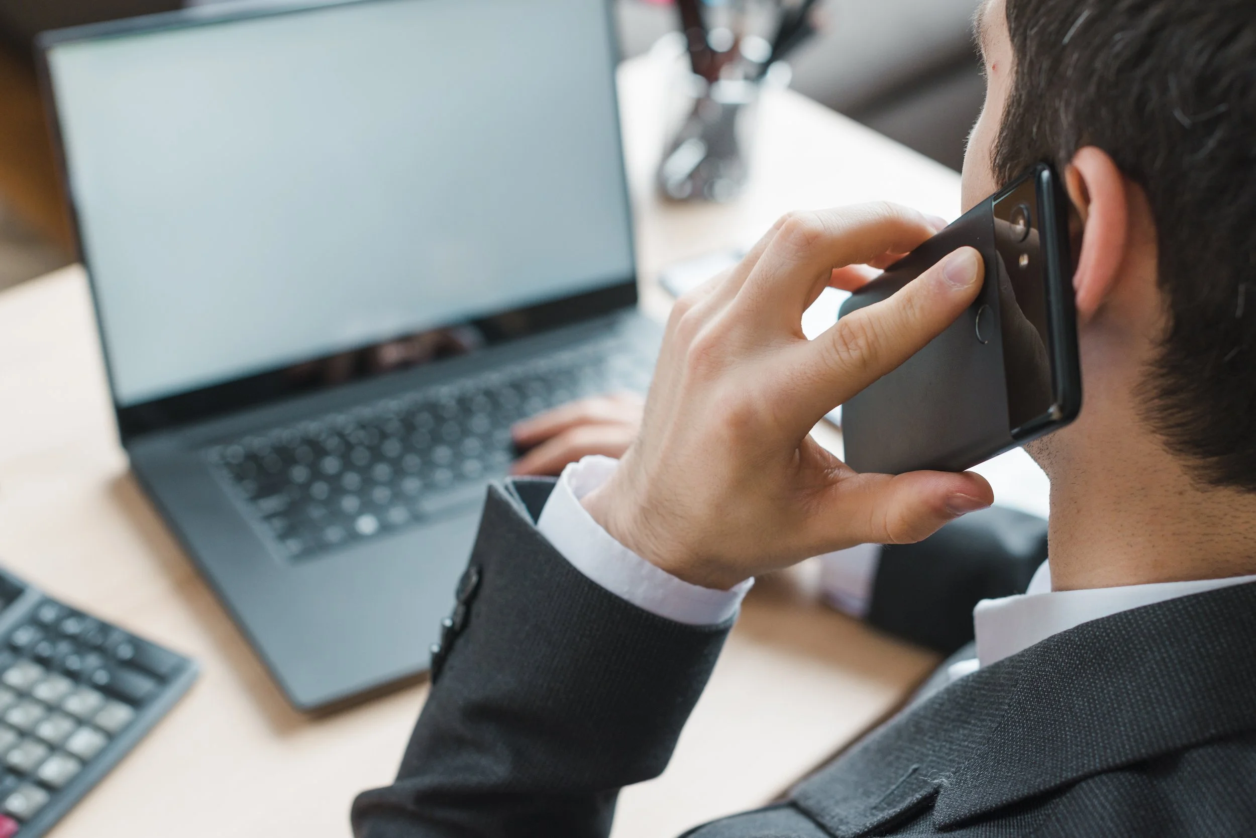 Man in business attire talking on smartphone at office desk with open laptop and calculator