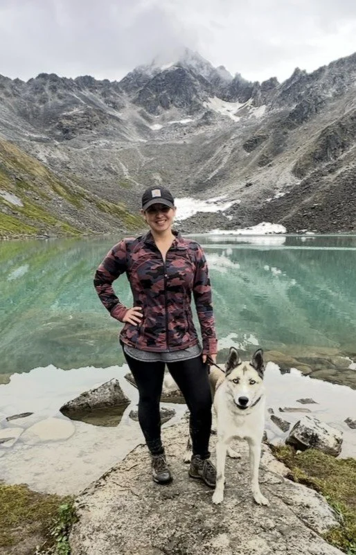 A woman and a Siberian Husky dog standing on rocks by a mountain lake with snowy peaks in the background.