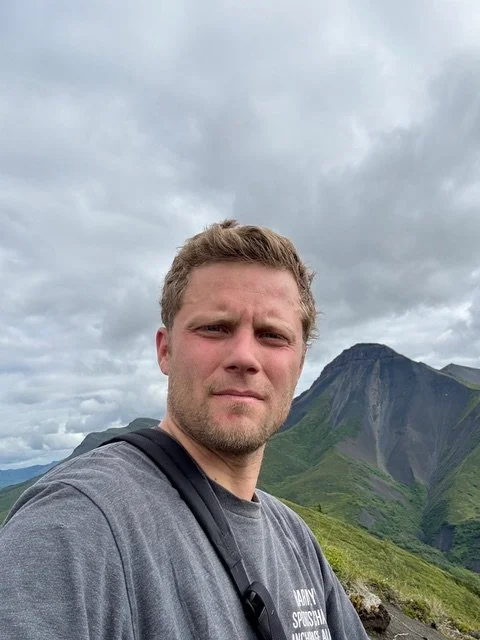 A young man taking a selfie outdoors with mountainous terrain and cloudy sky in the background.