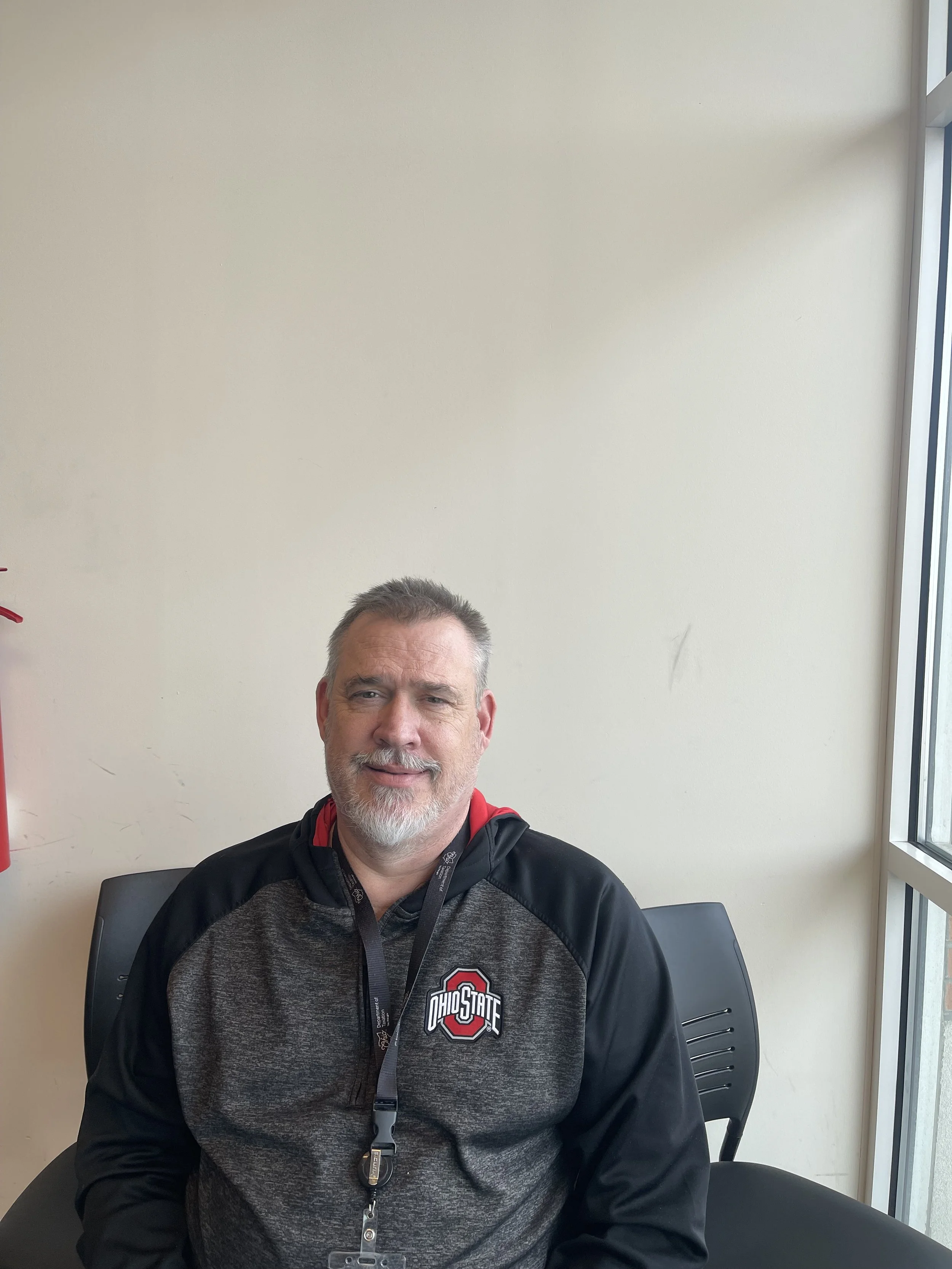A middle-aged man with black and gray hair sitting on a beige bench against a plain, softly lit wall