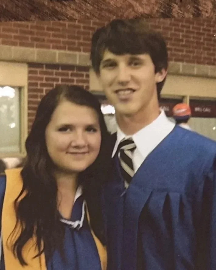 A young woman and a young man in graduation gowns and caps, smiling indoors with a brick wall behind them.