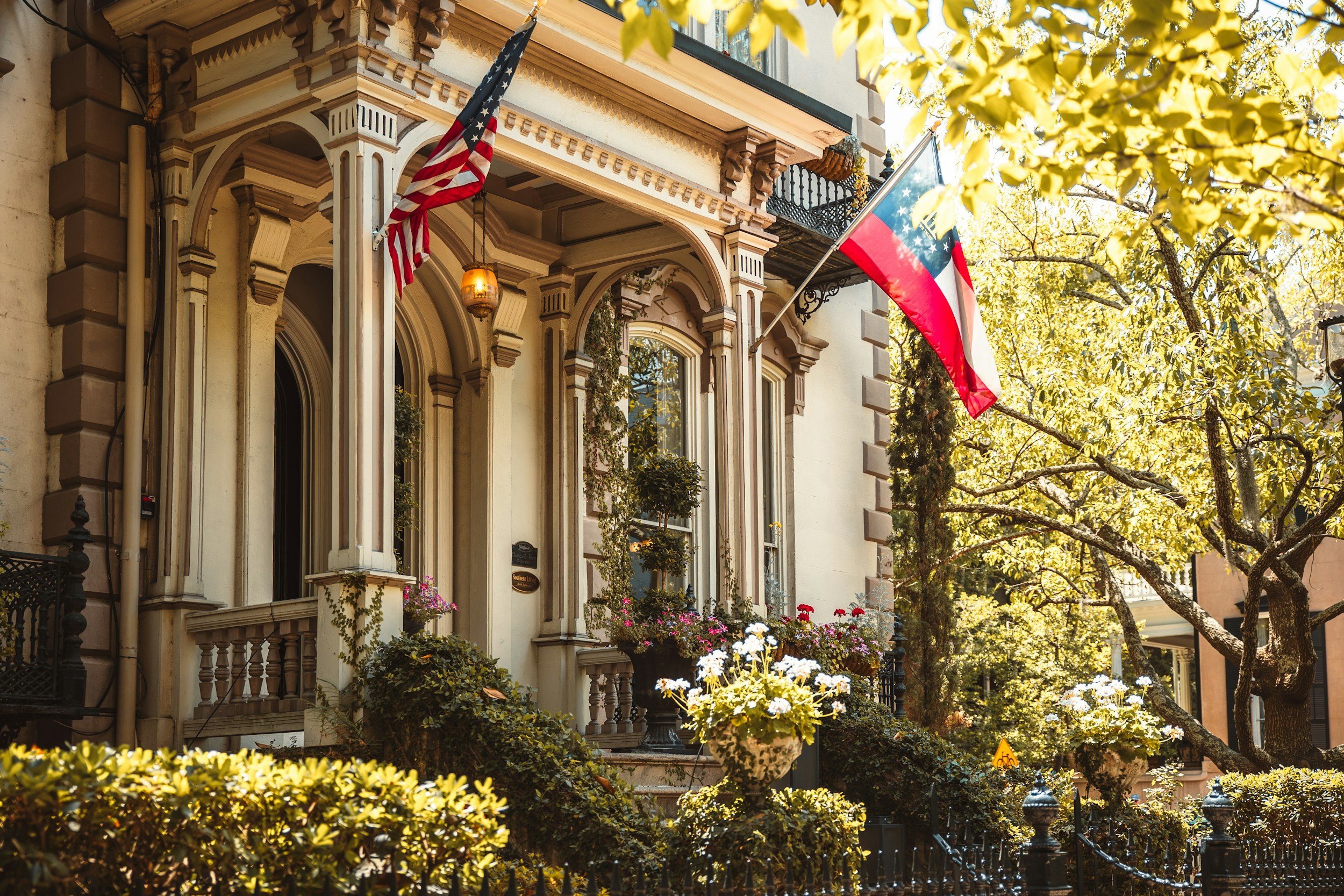 A historic residential building with ornate architecture, displaying American and Missouri state flags outside on a sunny day, surrounded by green trees and colorful flowers.