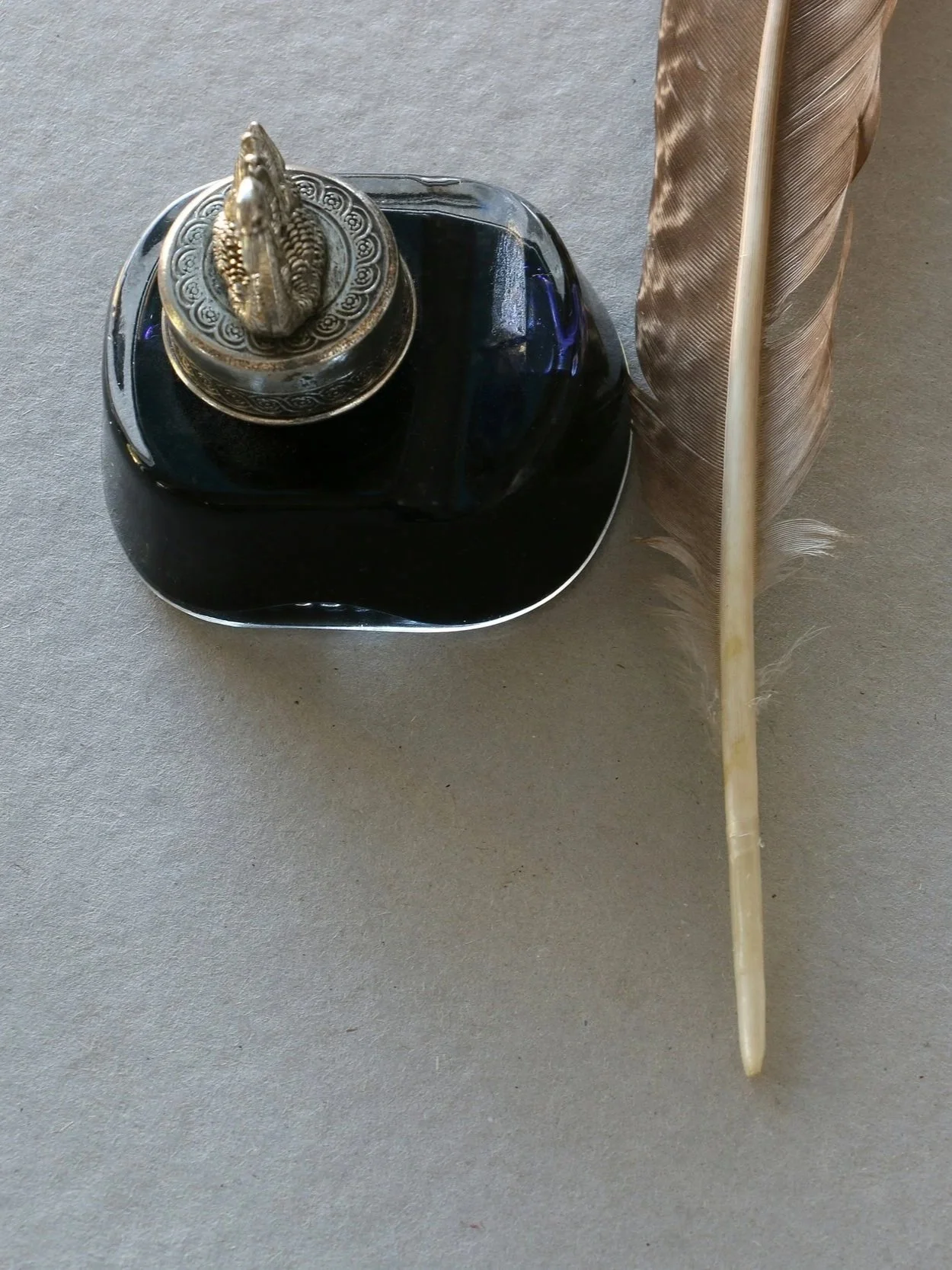 A feather, an ink bottle with a decorative metallic lid, and a feather quill pen on a flat surface.