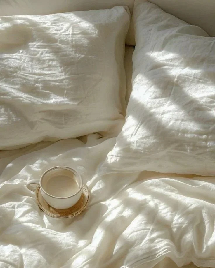 A cozy bed with white, slightly wrinkled sheets and two pillows. There is a cup of coffee or tea on a small, round wooden tray placed on the bed.