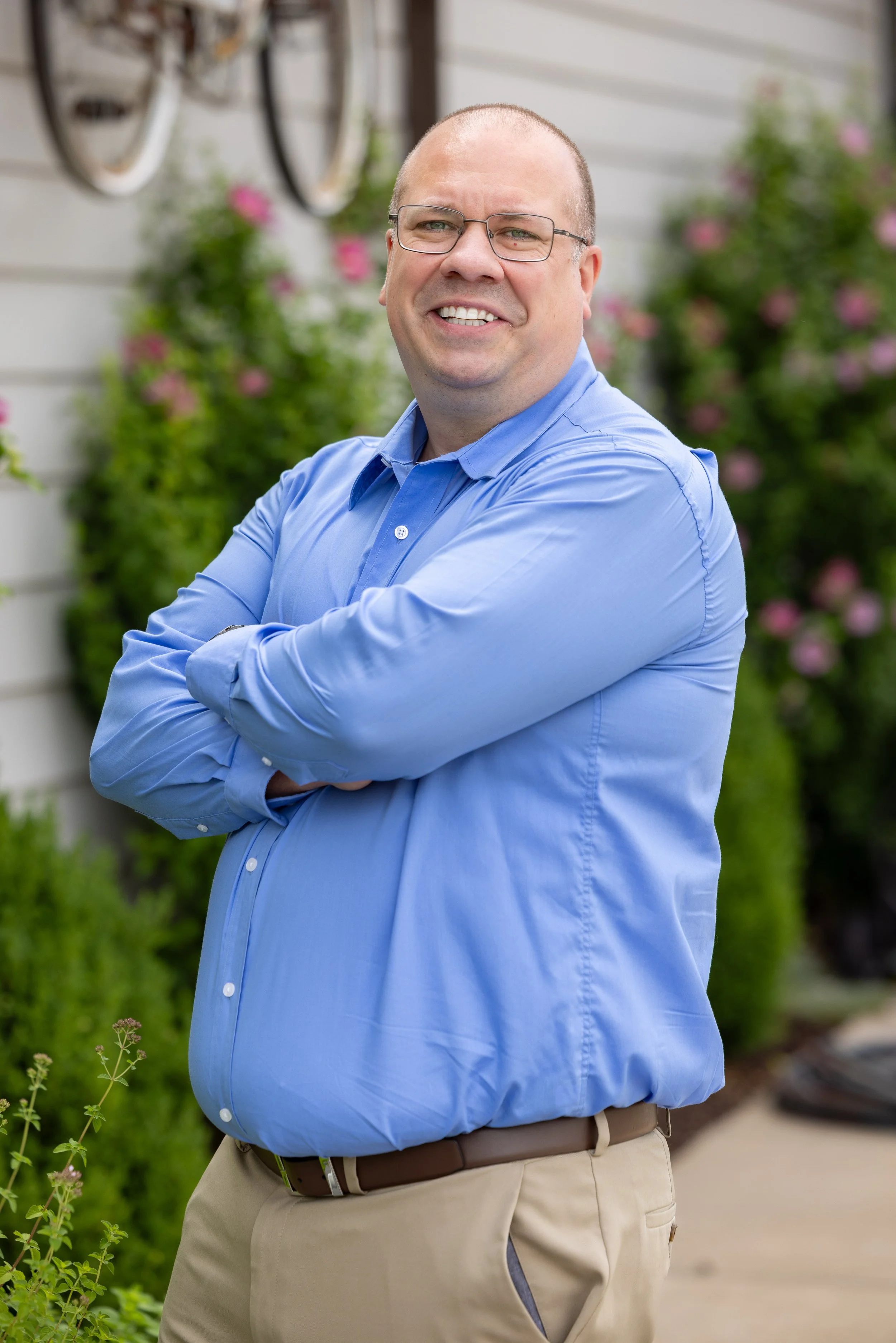 A man with glasses wearing a blue dress shirt and beige pants stands outdoors with his arms crossed, smiling. There are pink flowers and green bushes in the background.