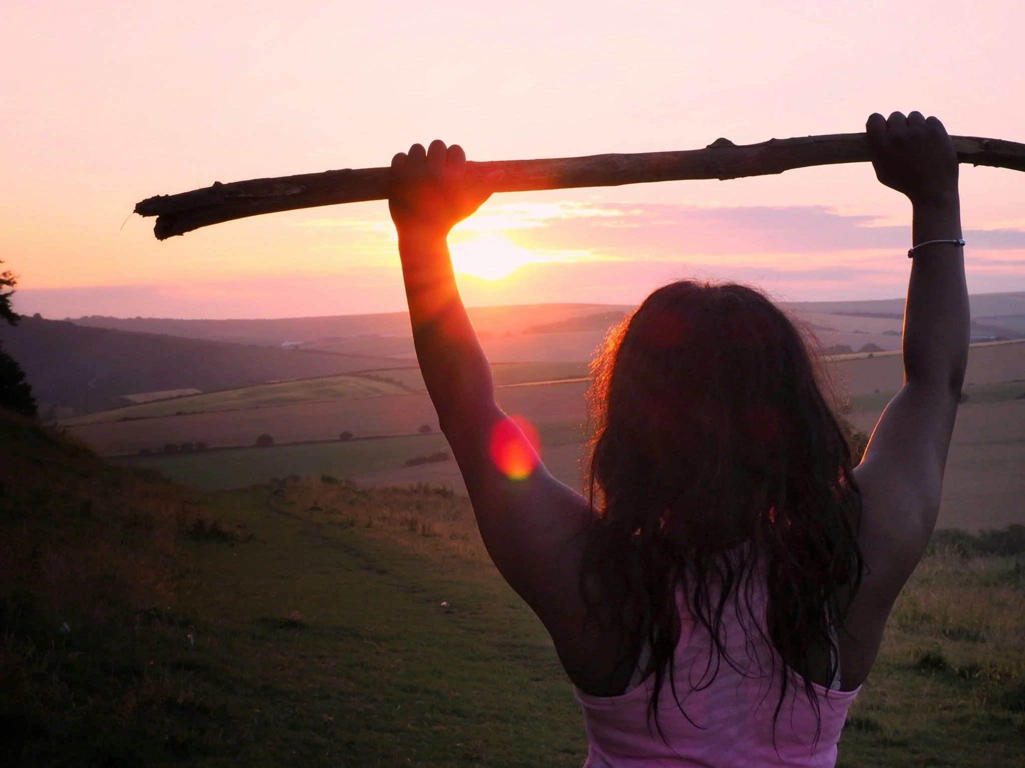 A woman with curly hair wearing a pink tank top stands outdoors during sunset, holding a large stick above her head with both hands, overlooking rolling hills and fields.