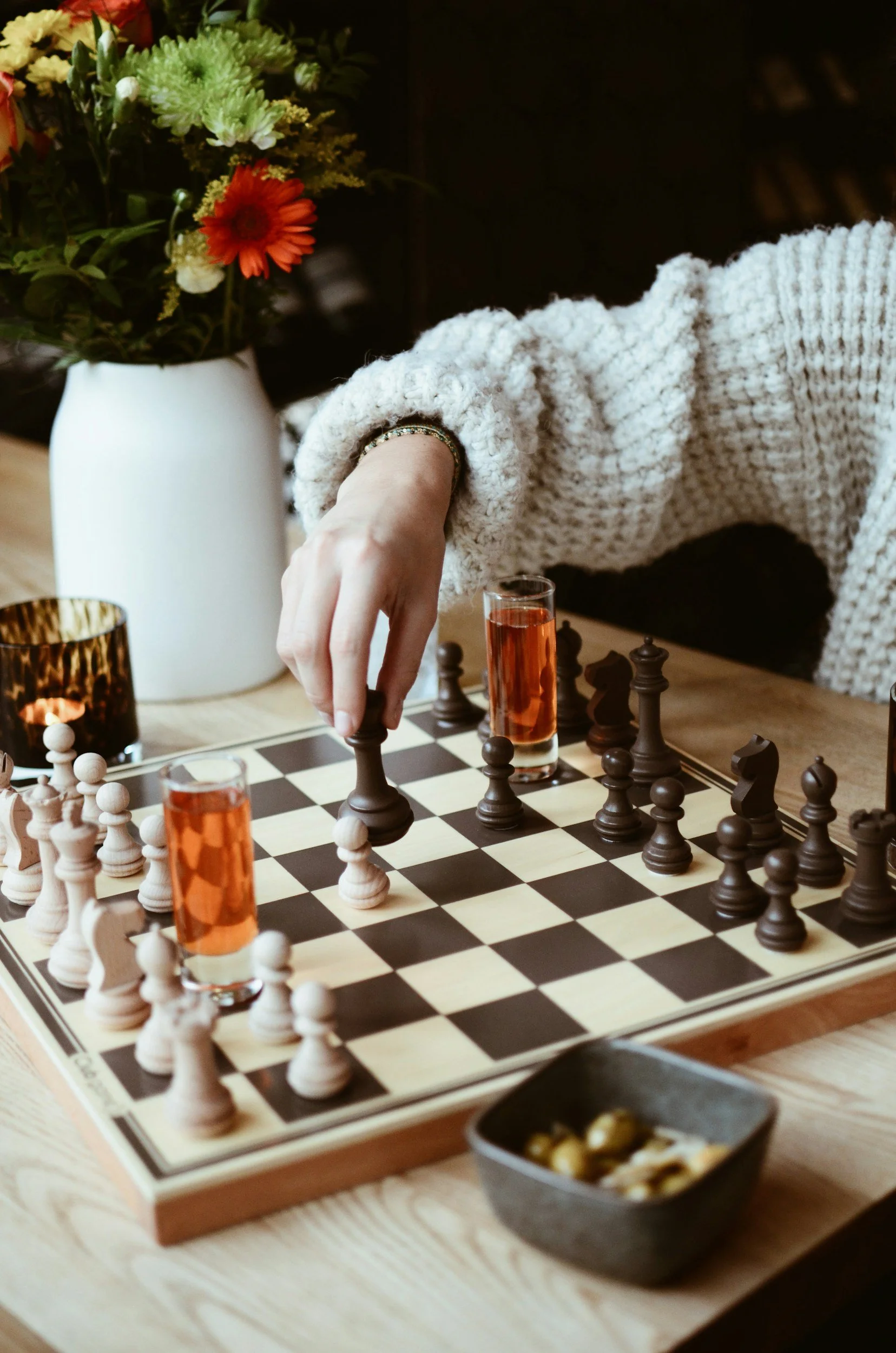 A person in a cream-colored knitted sweater moving a black chess piece on a chessboard. Two glasses with a pink beverage are placed on the chessboard. There is a large white vase with colorful flowers and a small black bowl with snacks on the wooden table.