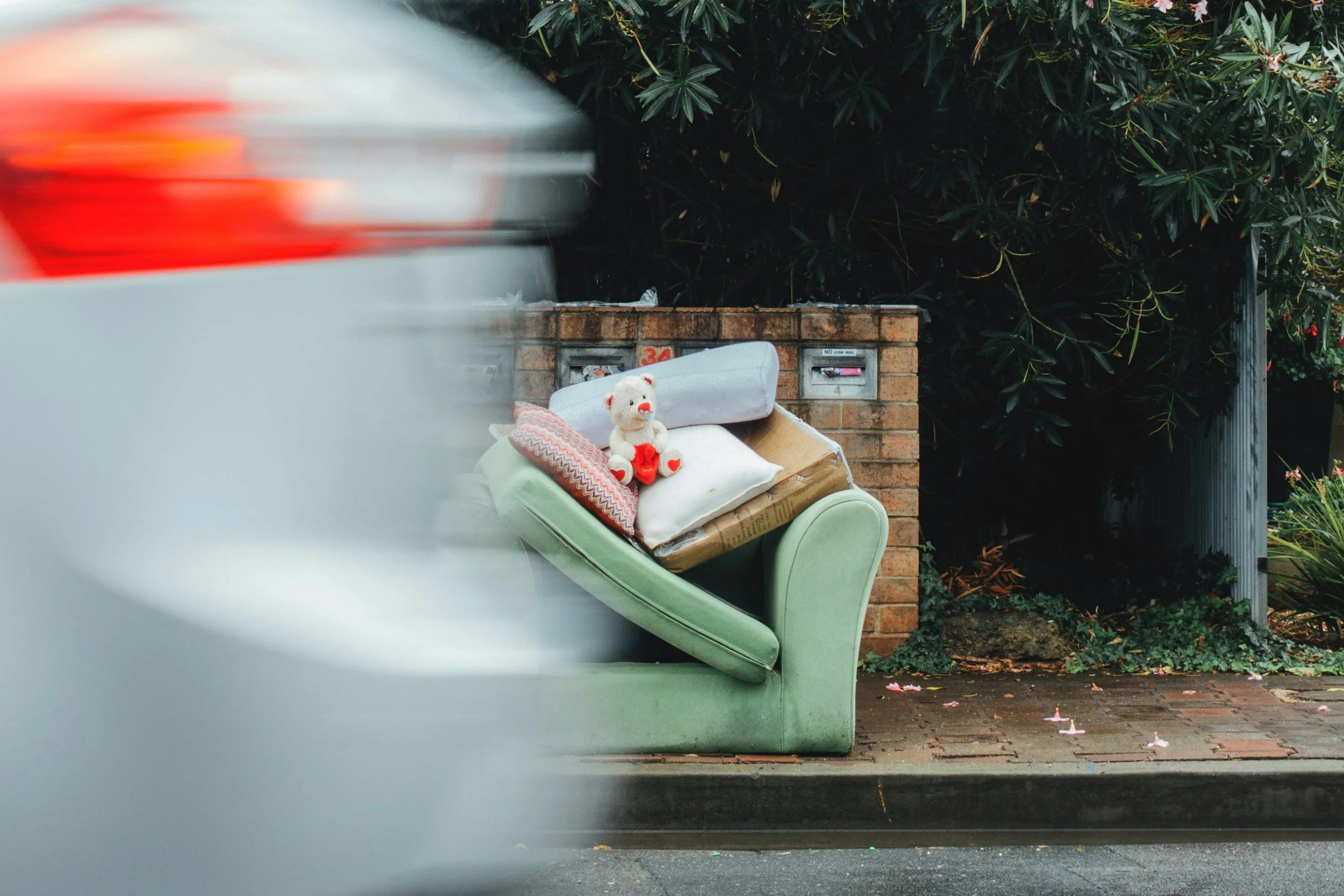 An abandoned green armchair filled with pillows and a teddy bear, placed next to a brick wall with mailboxes, on a sidewalk with fallen petals.