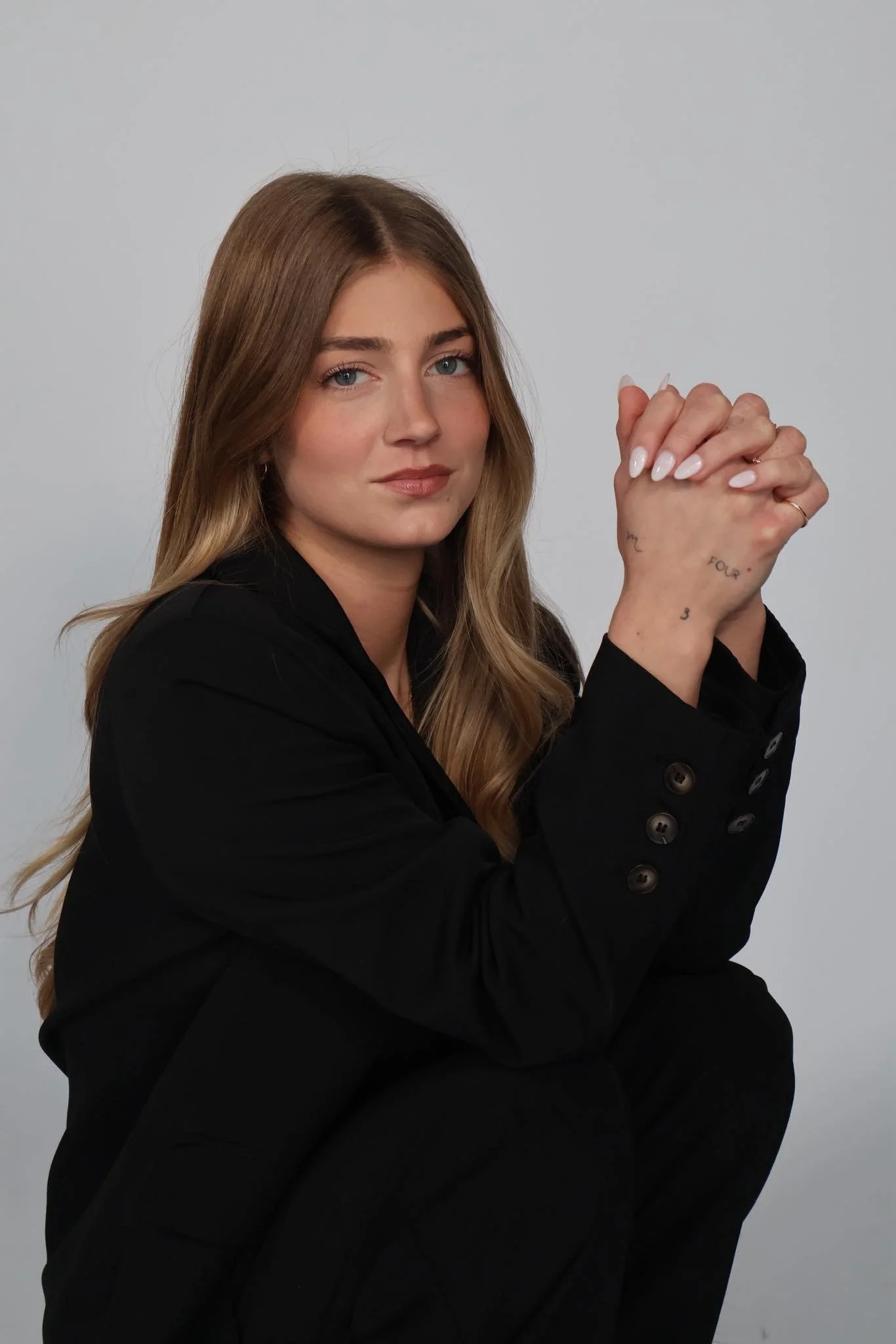 A woman with long, wavy, light brown hair and blue eyes, wearing a black blazer, poses with her hands clasped, showing tattoos on her fingers and wrist, against a plain background.