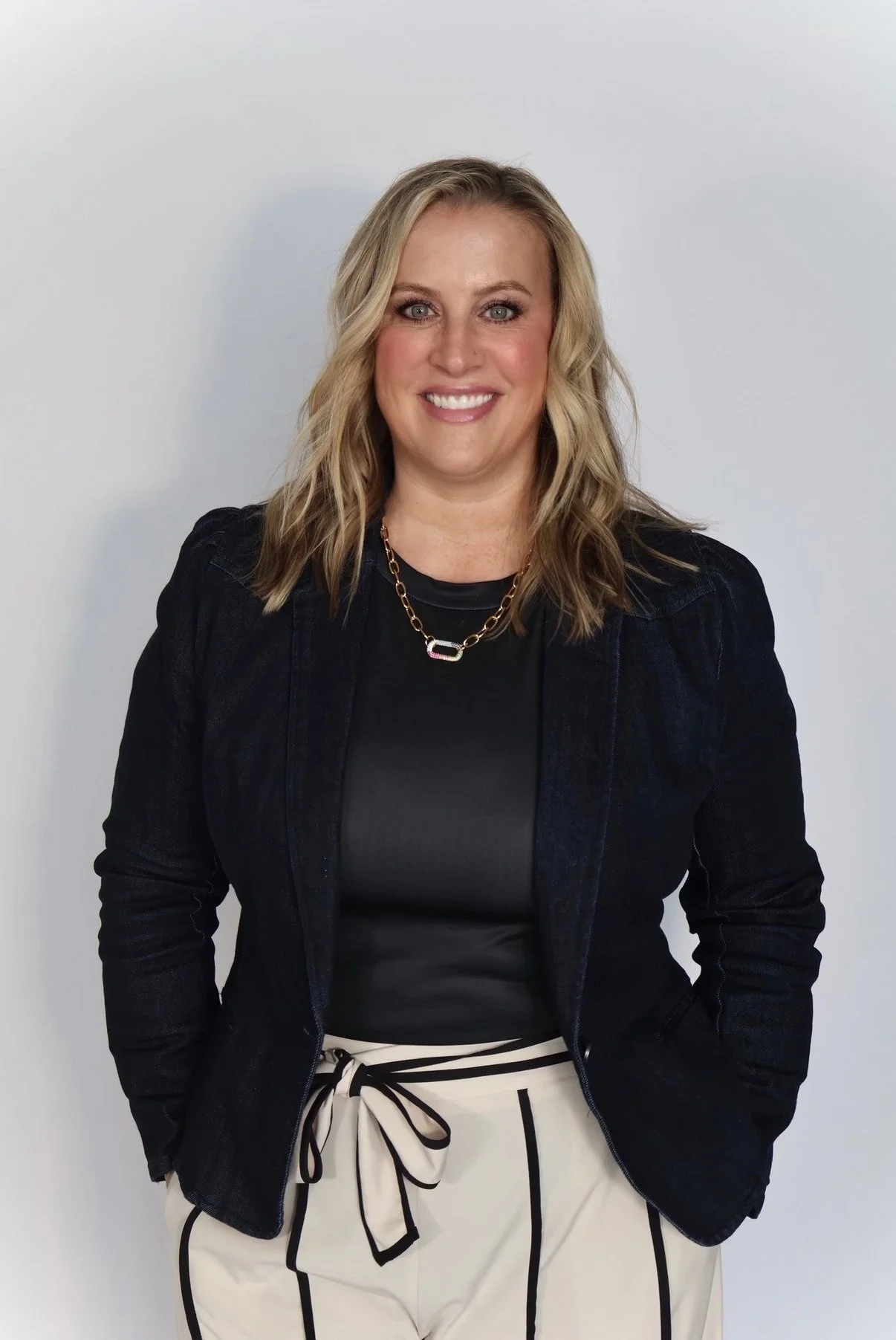 A woman with blonde wavy hair smiling, wearing a black top, black blazer, and white pants with black stripes and a bow, standing against a light gray background.