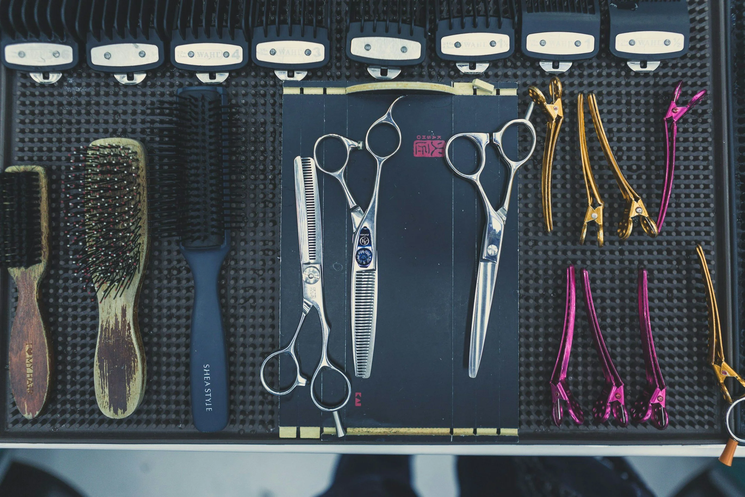 Hairdressing tools including brushes, scissors, and hair clips arranged on a black textured mat.