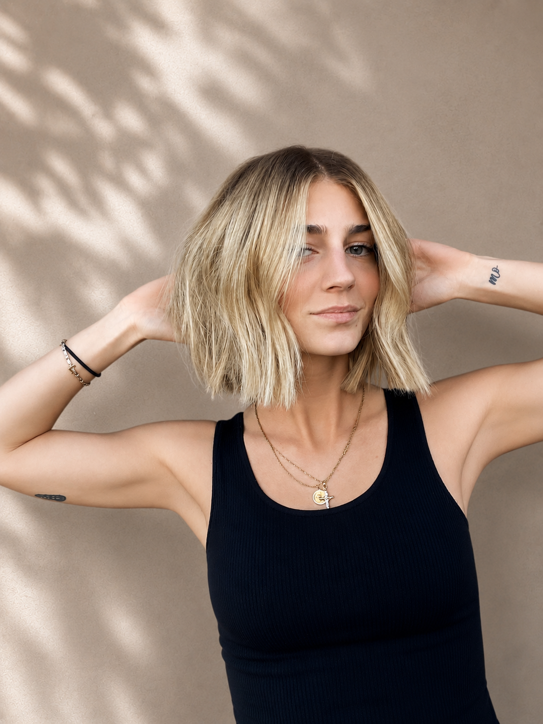 A young woman with shoulder-length blonde hair and fair skin is posing with her hands behind her head, wearing a black tank top and layered necklaces, against a neutral beige background with soft shadows.