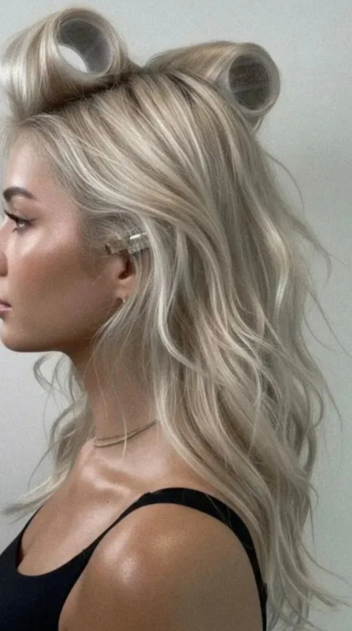 Side profile of a woman with platinum blonde hair in large curlers, wearing a black tank top and a thin necklace, standing against a plain wall.